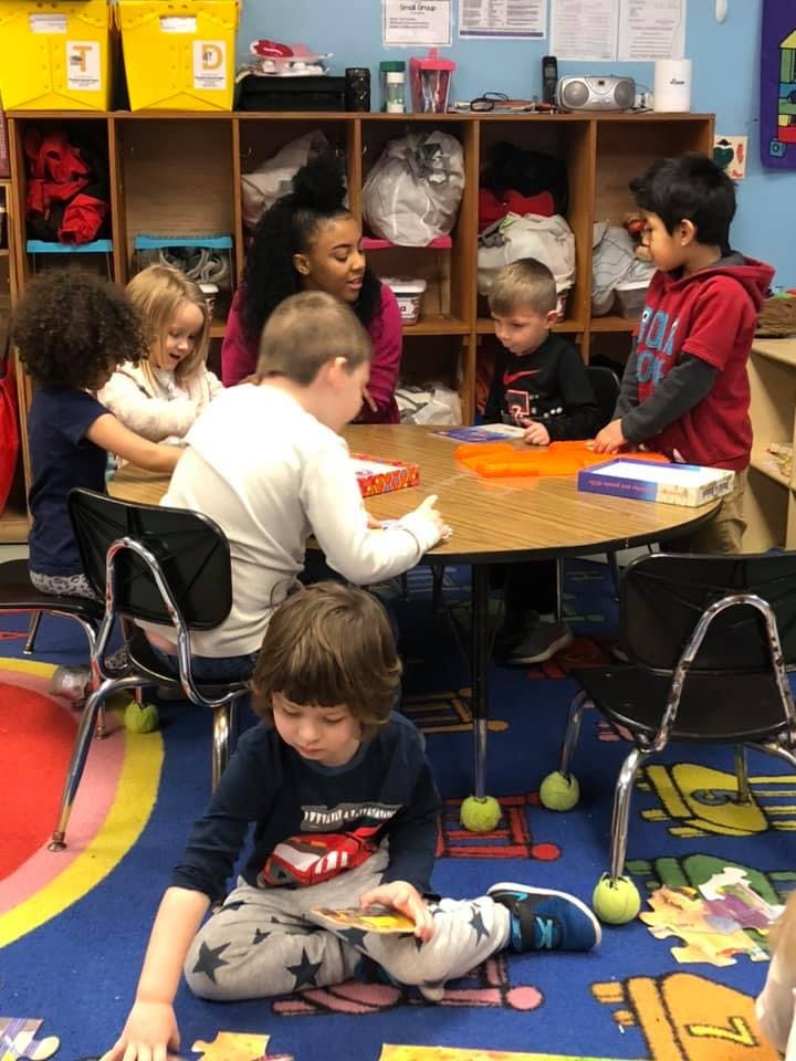 Adult and children sitting at a table and on the floor in a classroom