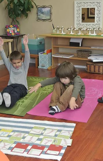 Two children sitting on yoga mats, one with arms raised