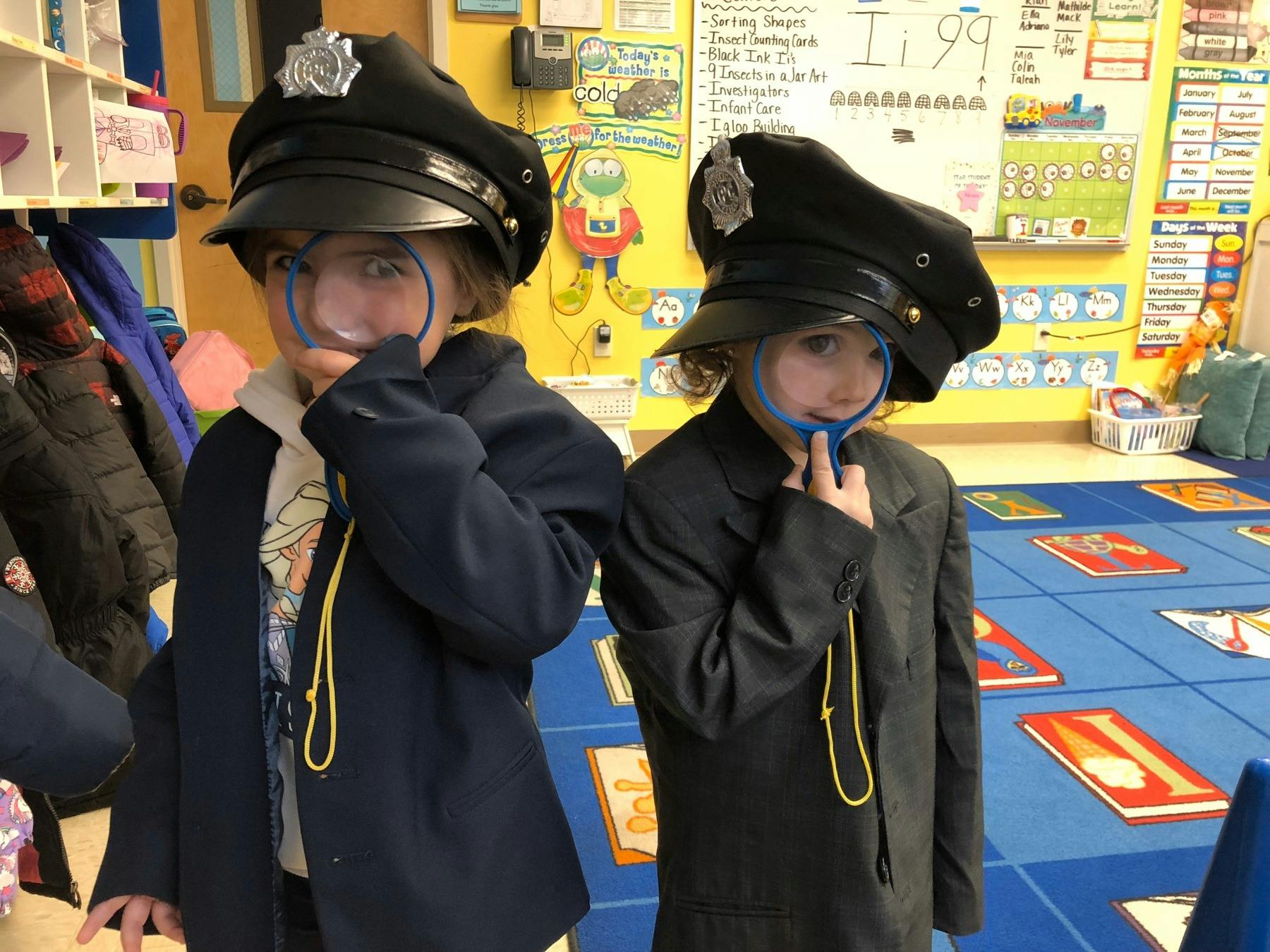 Two children in police hats and coats holding magnifying glasses in a classroom