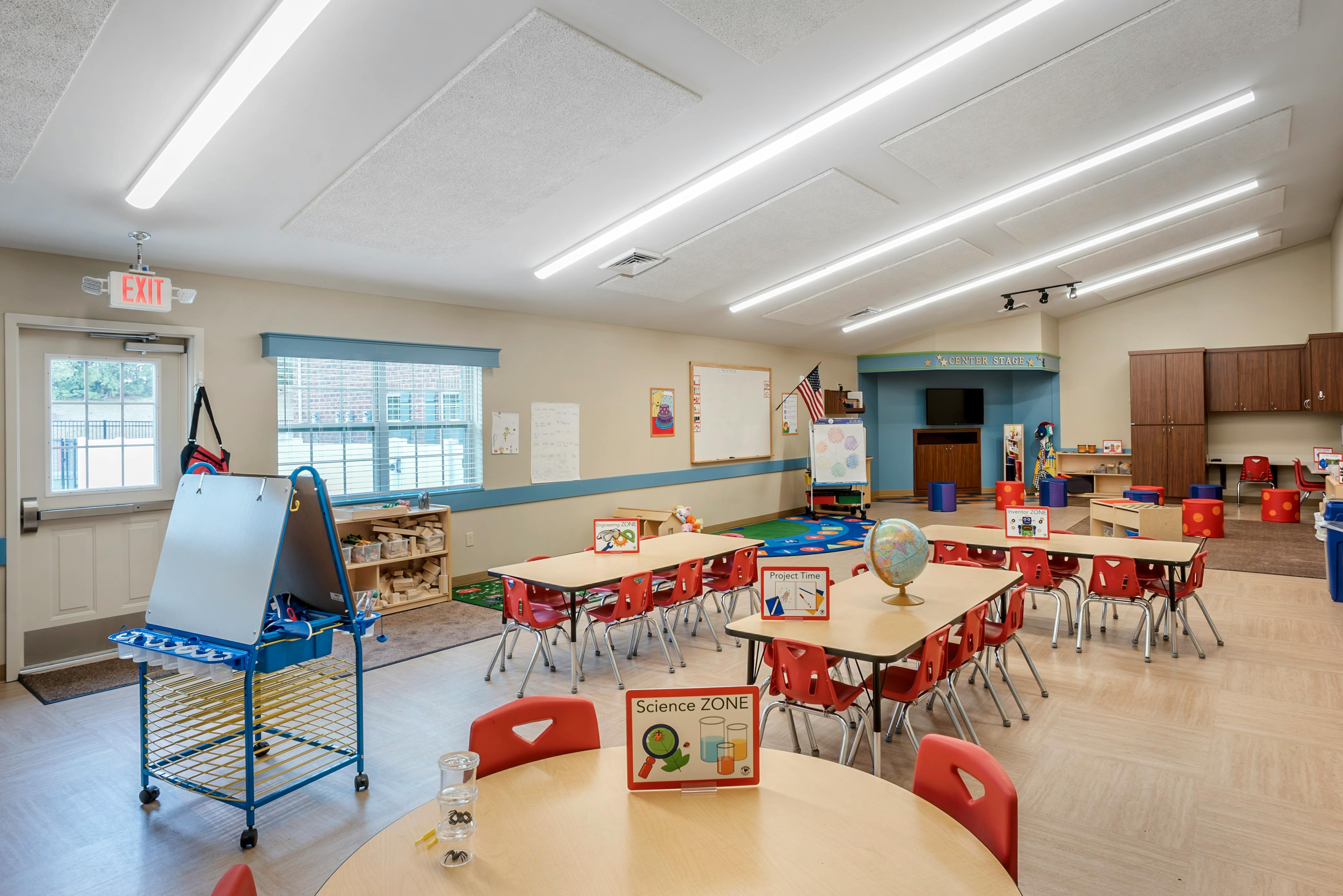 A classroom with small tables and red chairs, an easel, and various learning zones