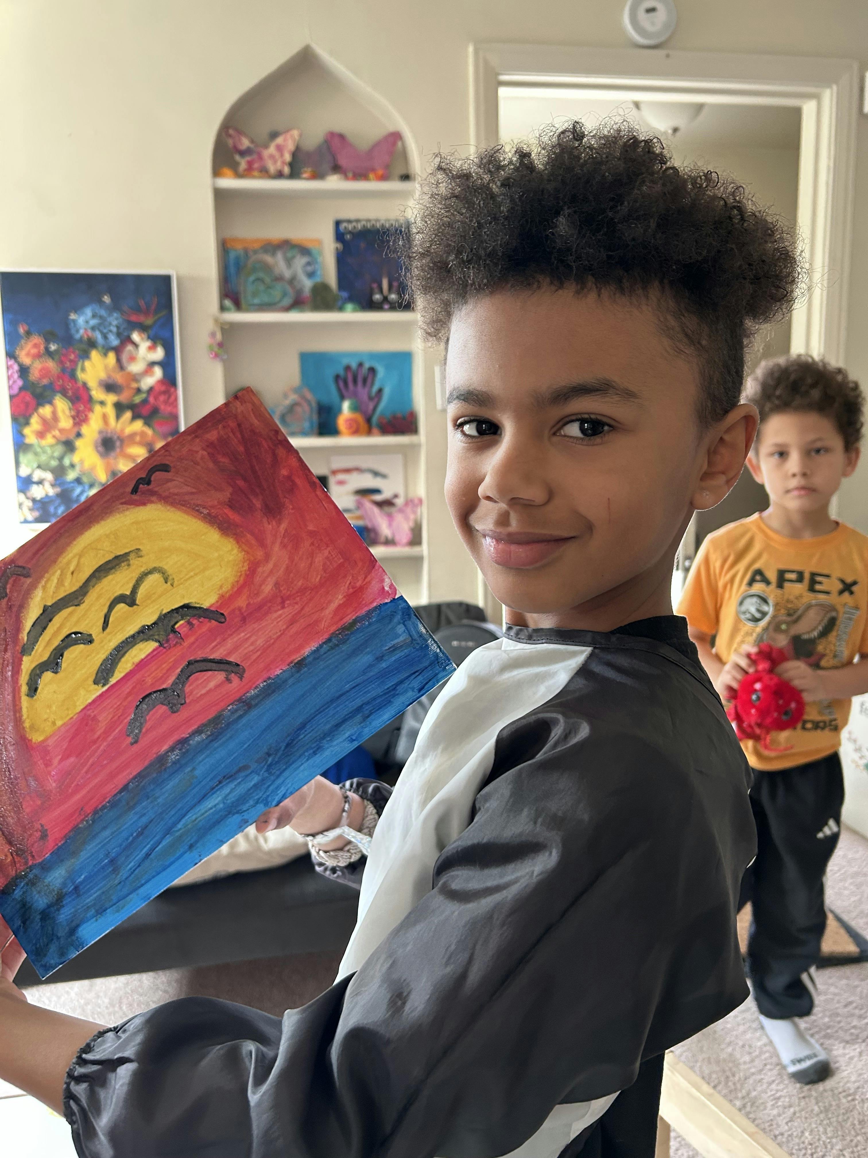 Child in smock holding up a painting of a sunset over water with birds flying