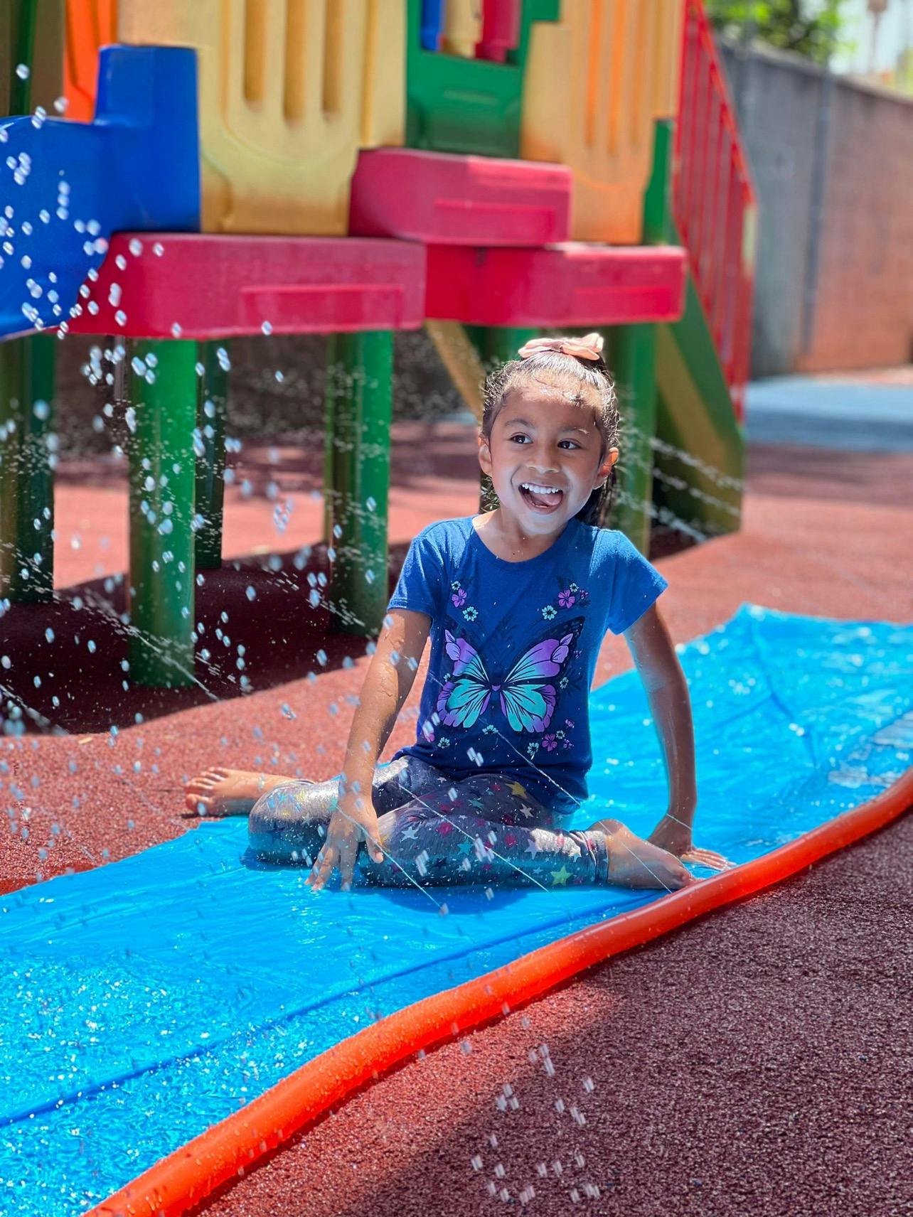 Child sitting on a blue slip-n-slide with water spraying, smiling