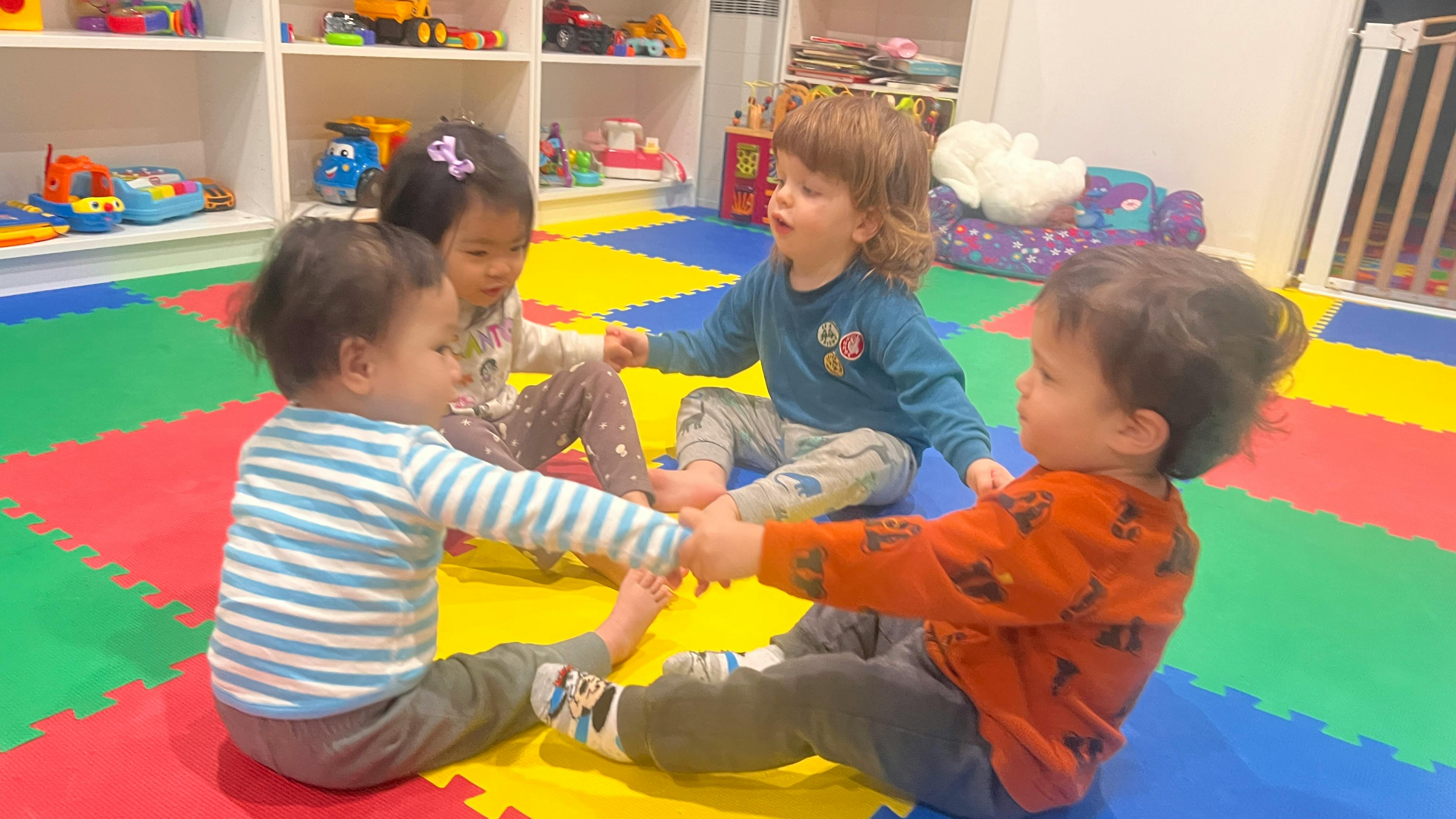 Four children sitting in a circle holding hands on a colorful mat