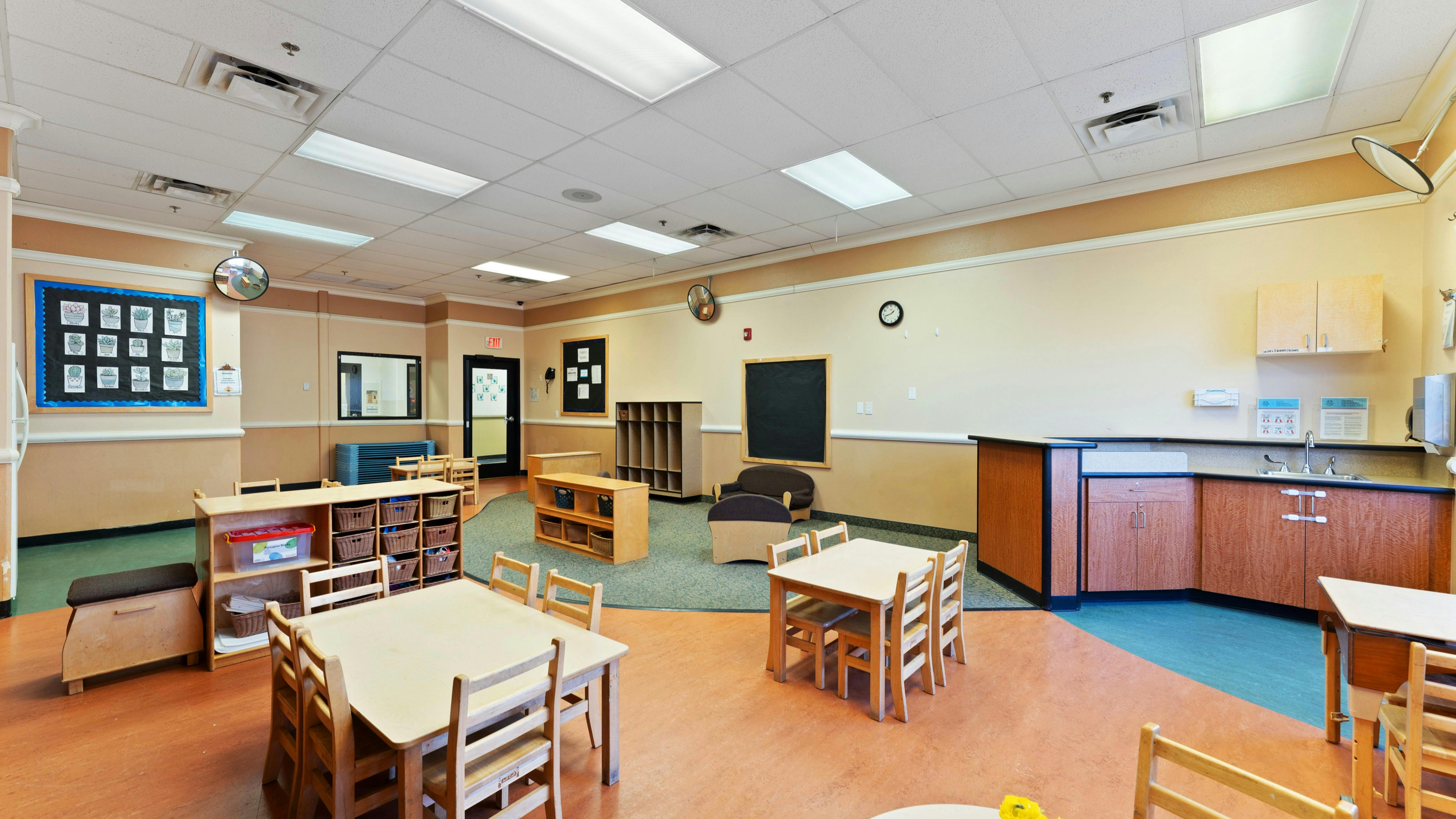 An empty classroom with small tables and chairs, shelves, and a sink area