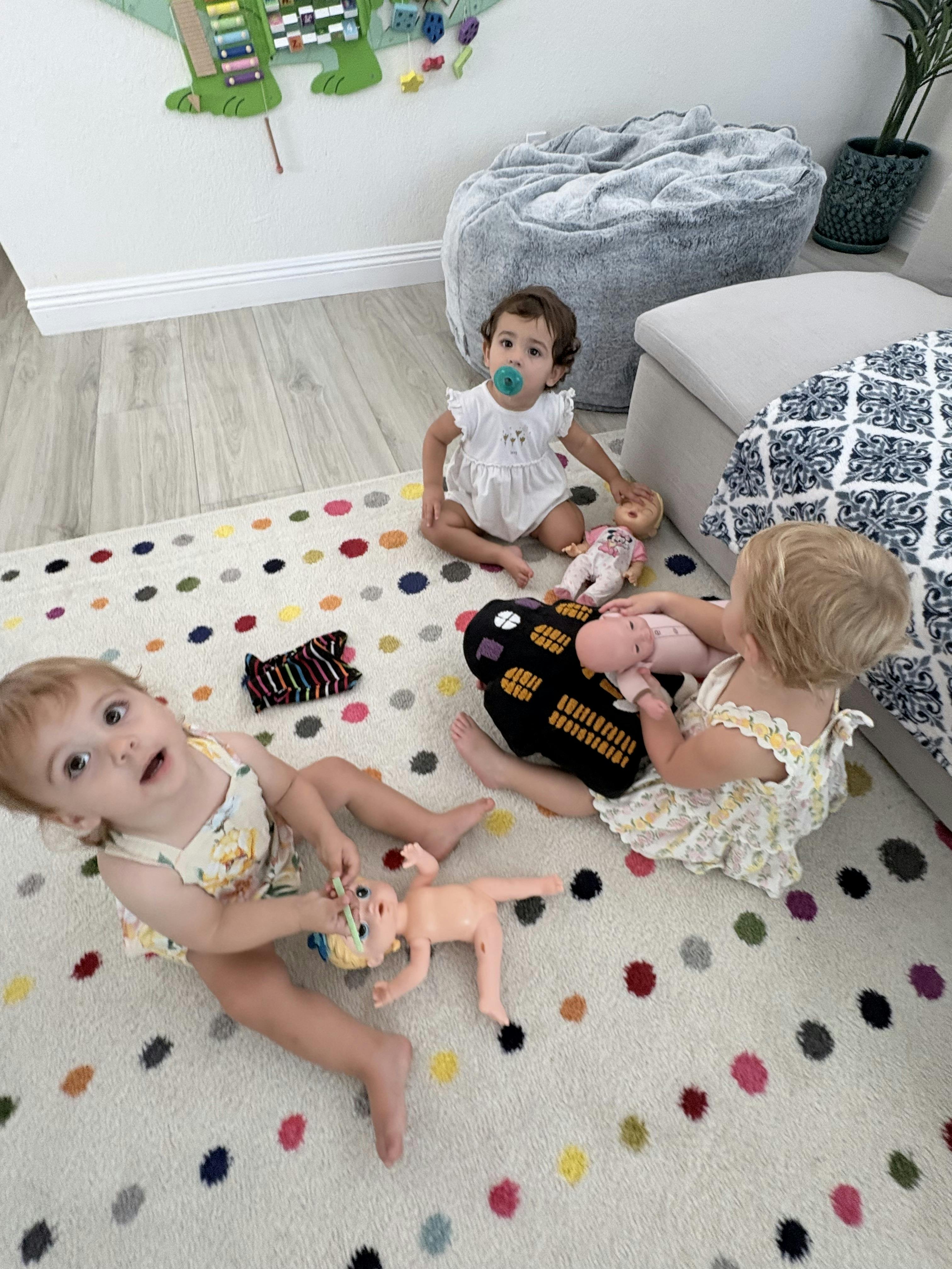 Three children playing with dolls on a polka dot rug in a playroom