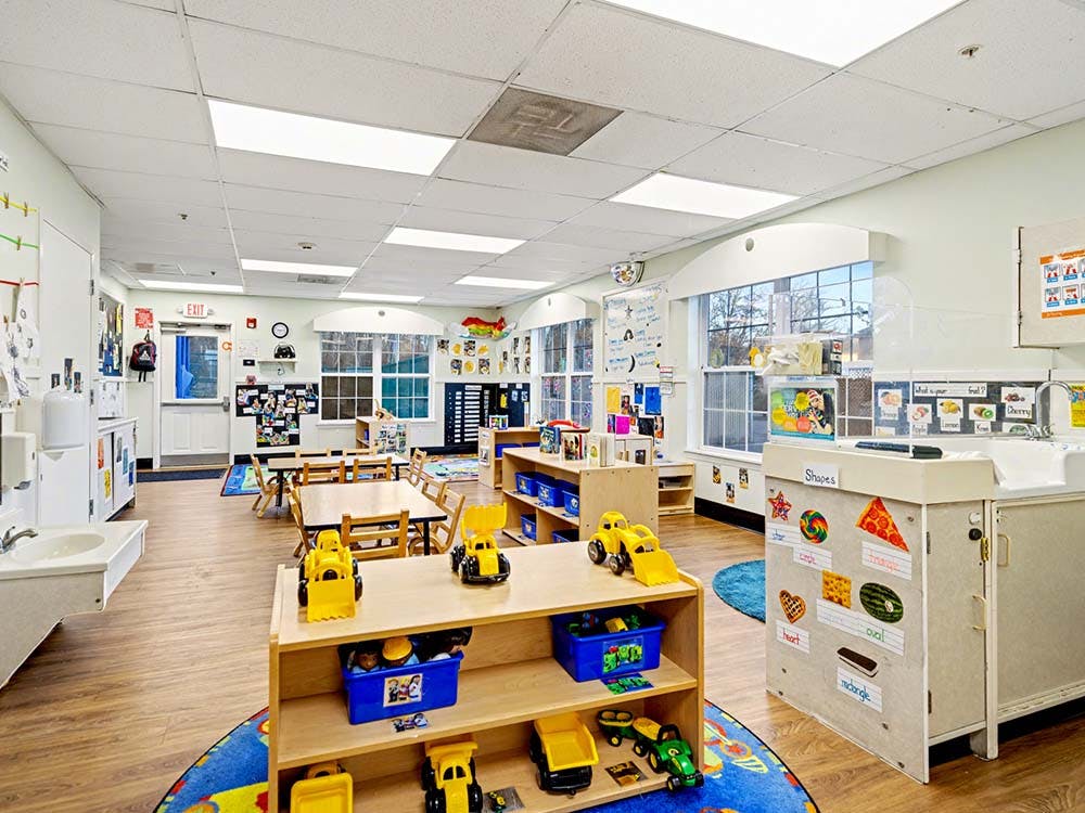 Empty classroom with small tables and chairs, shelves with toys, and posters