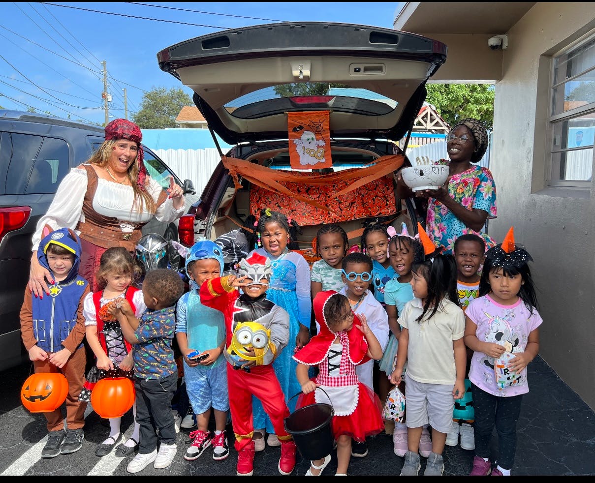 Two adults and many children in costumes standing by an open car trunk