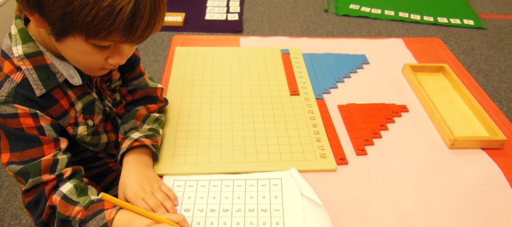 Child writing on paper with a pencil, next to math learning materials