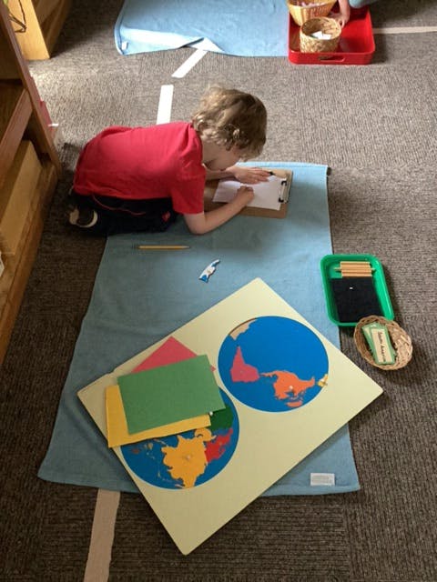 Child writing on a clipboard on the floor next to a world map puzzle