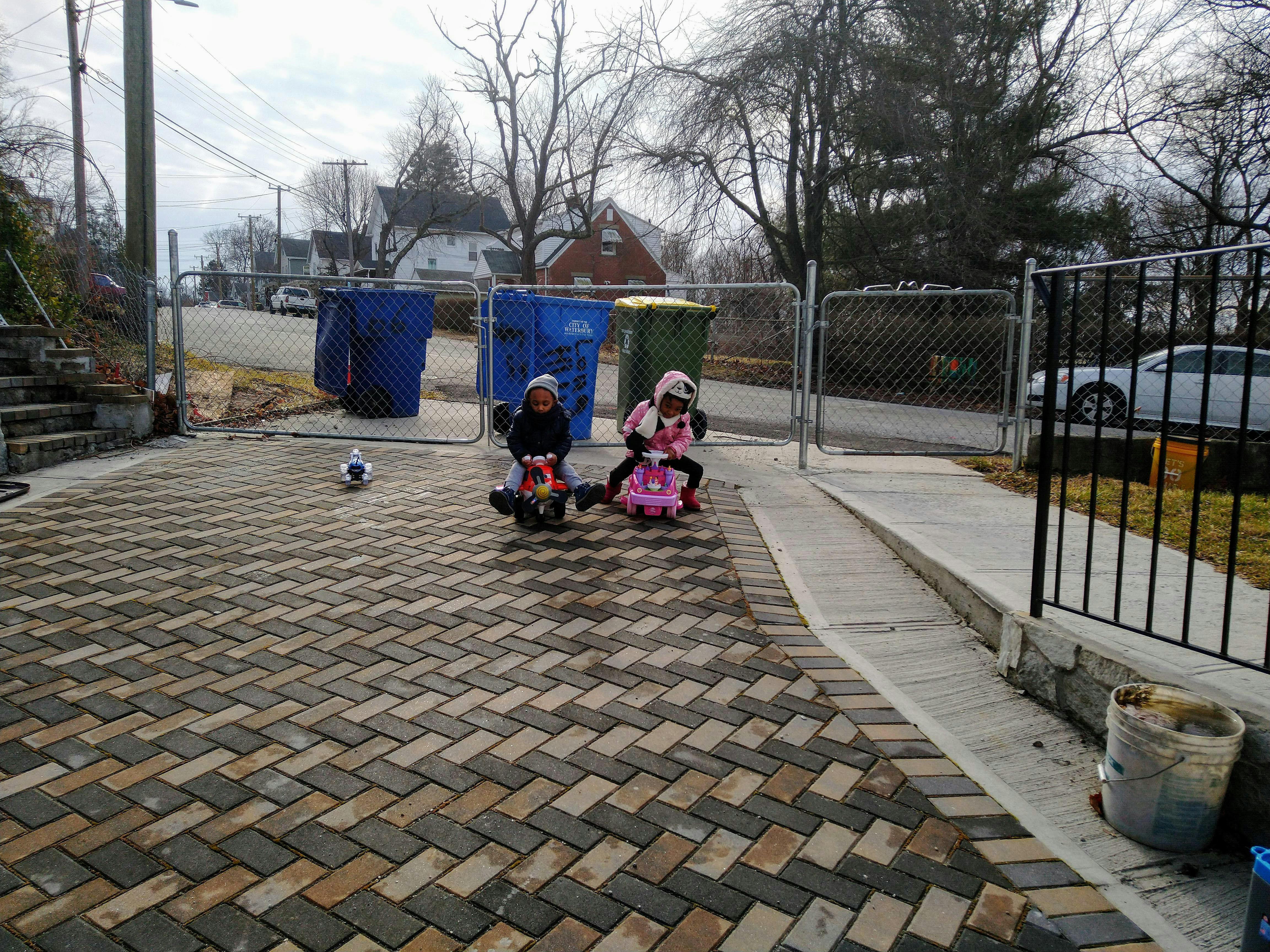 Two children riding toy cars on a paved driveway