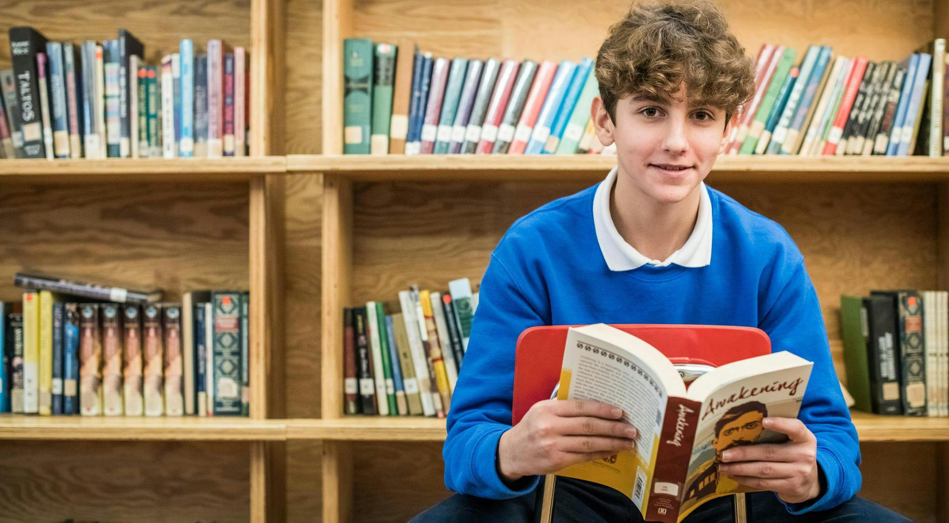 teen in a blue shirt holding an open book in front of bookshelves