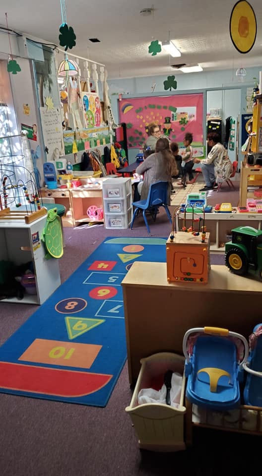 Adults and children in a classroom with toys and a number rug