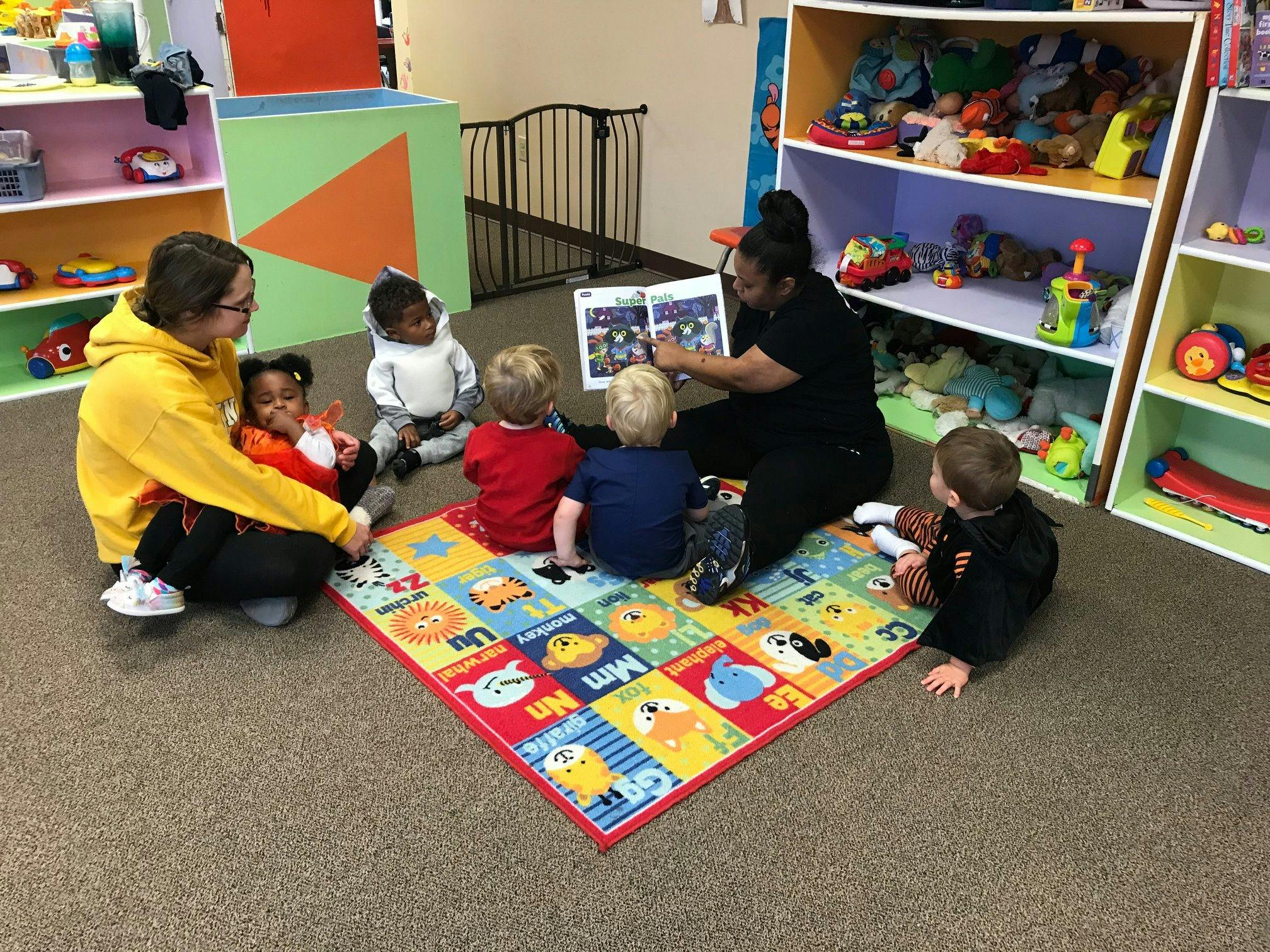 Two people reading to multiple children on a colorful mat in a classroom