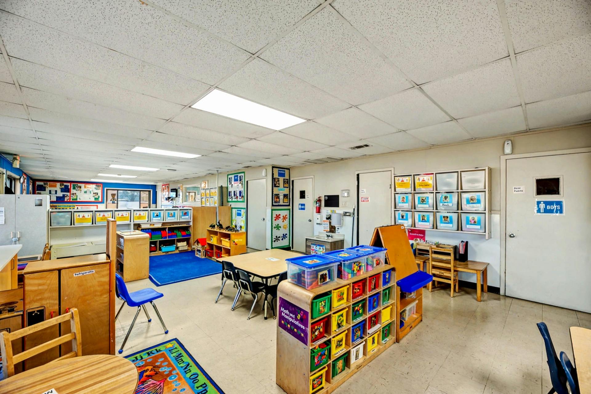 An empty classroom with small tables and chairs, shelves with toys and books