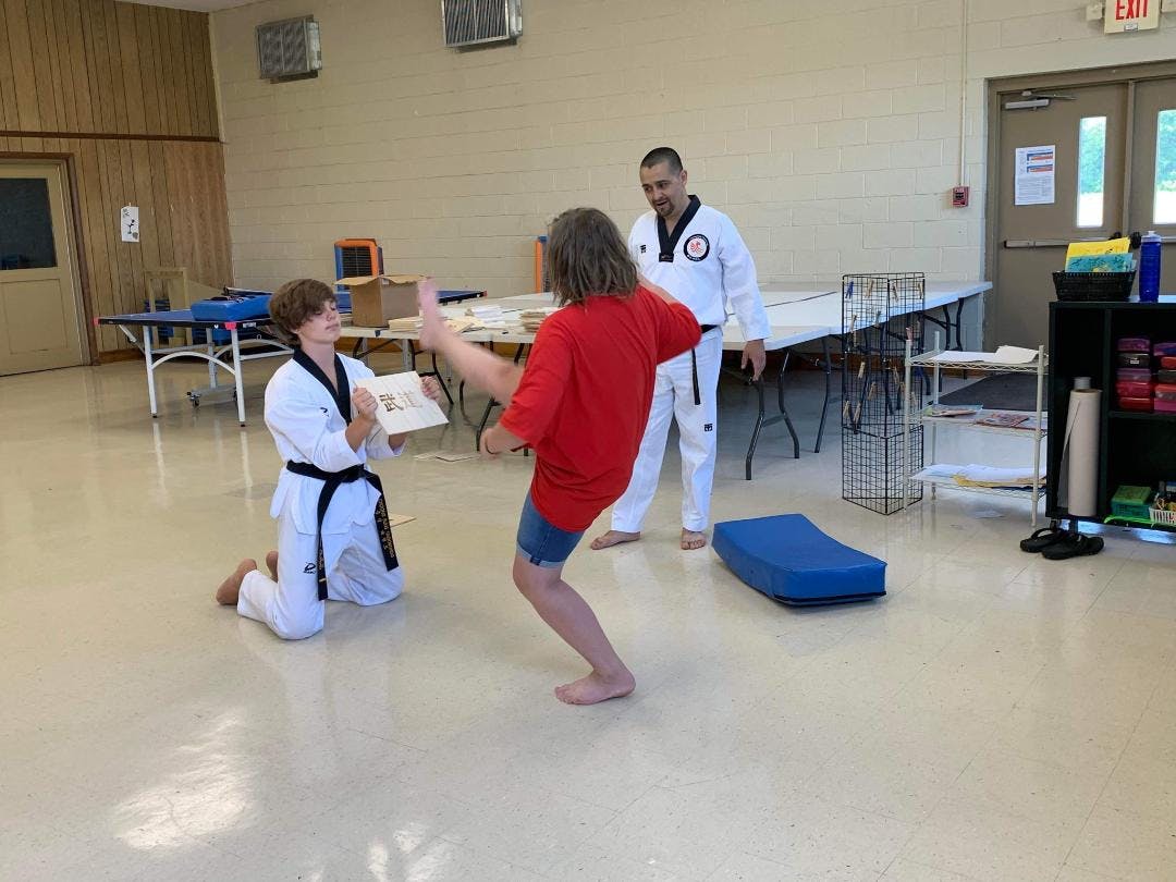 Person in taekwondo uniform kneeling, person in red shirt kicking, person in uniform watching