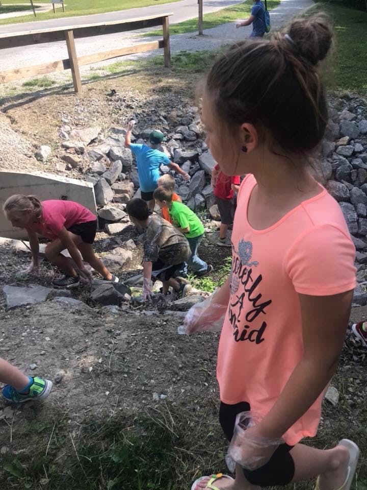 Child in pink shirt with other children picking up debris outdoors