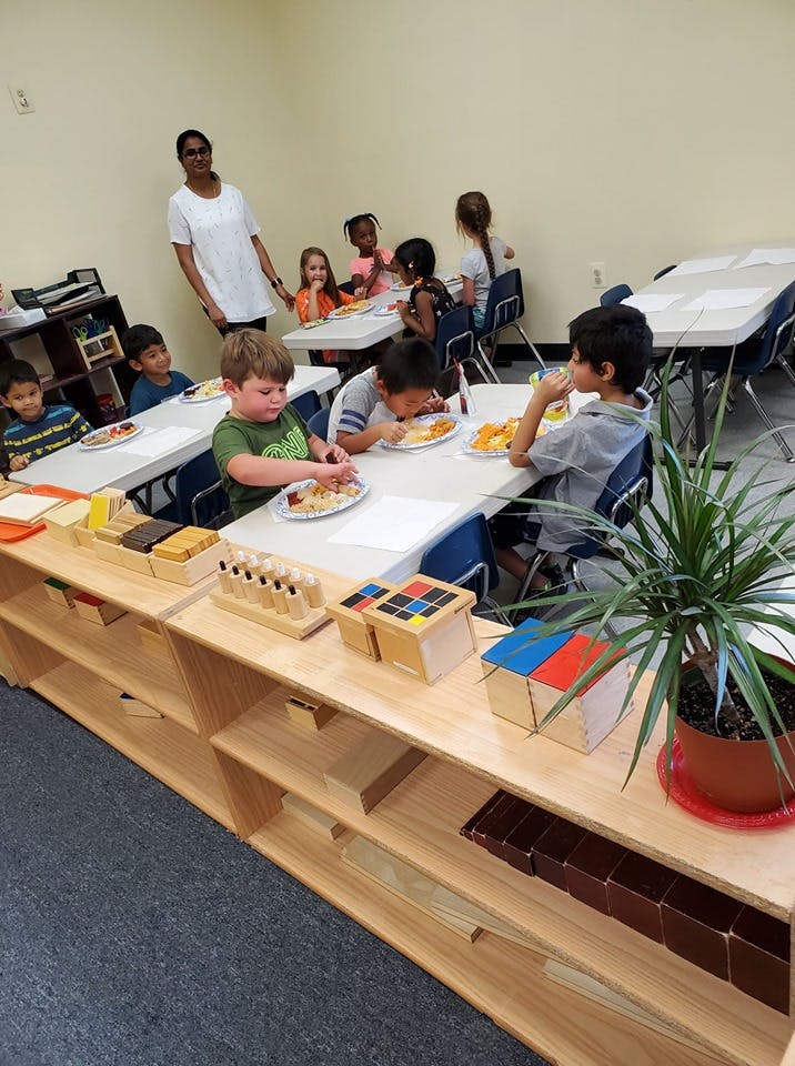 Children eating at tables in a classroom with an adult standing nearby