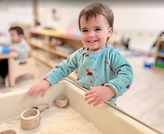 Child smiling while standing at a sensory bin with rice in a classroom