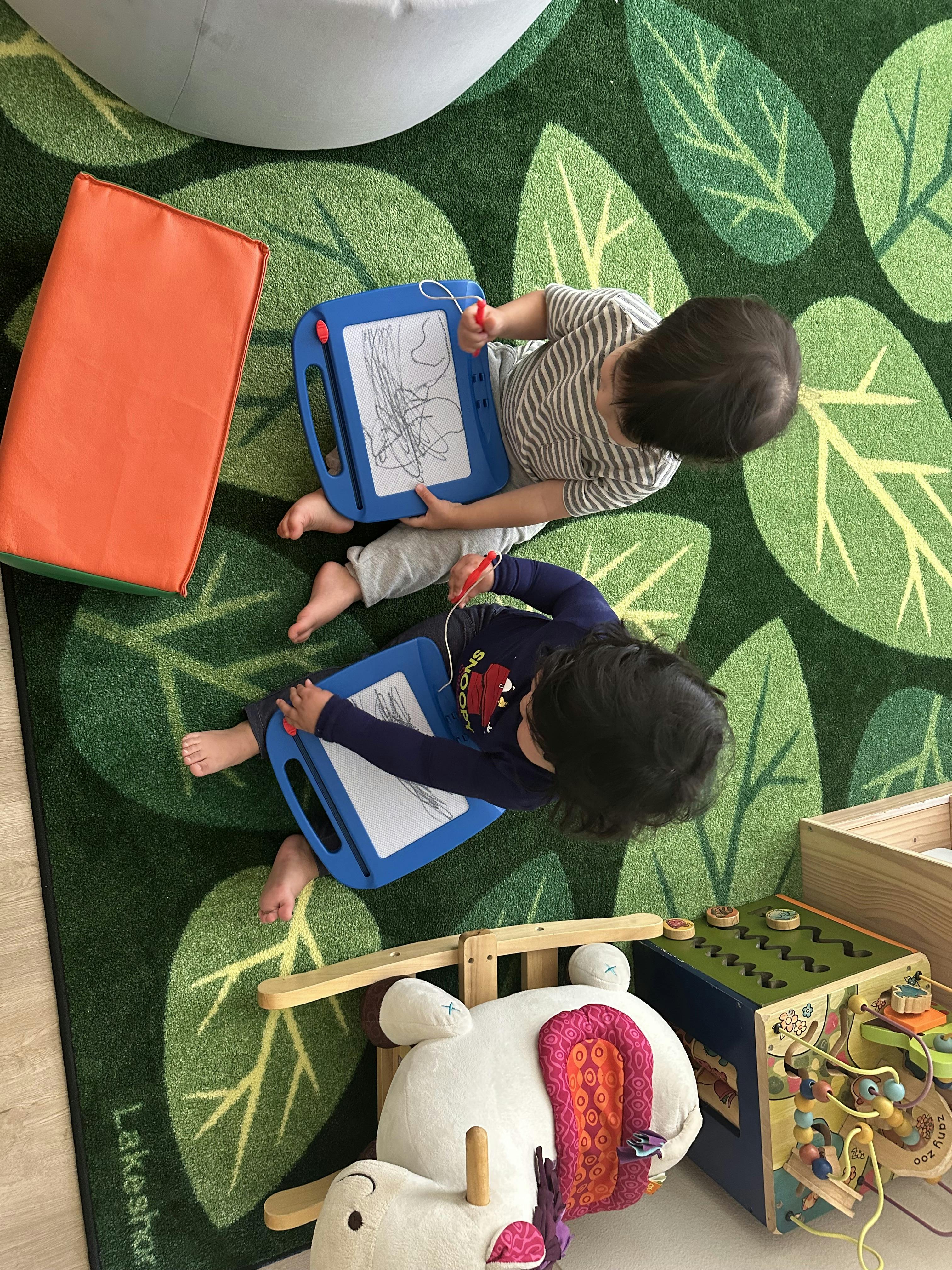 Two children sitting on a green leaf rug drawing on magnetic sketch boards