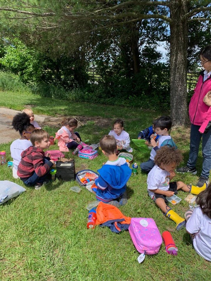 Children sitting on grass with lunchboxes and an adult standing nearby