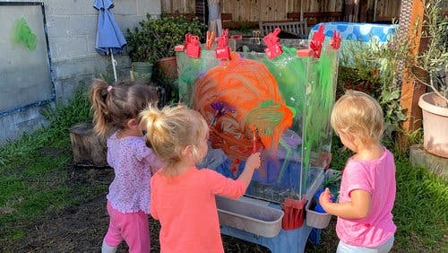 Three children painting on an easel outdoors