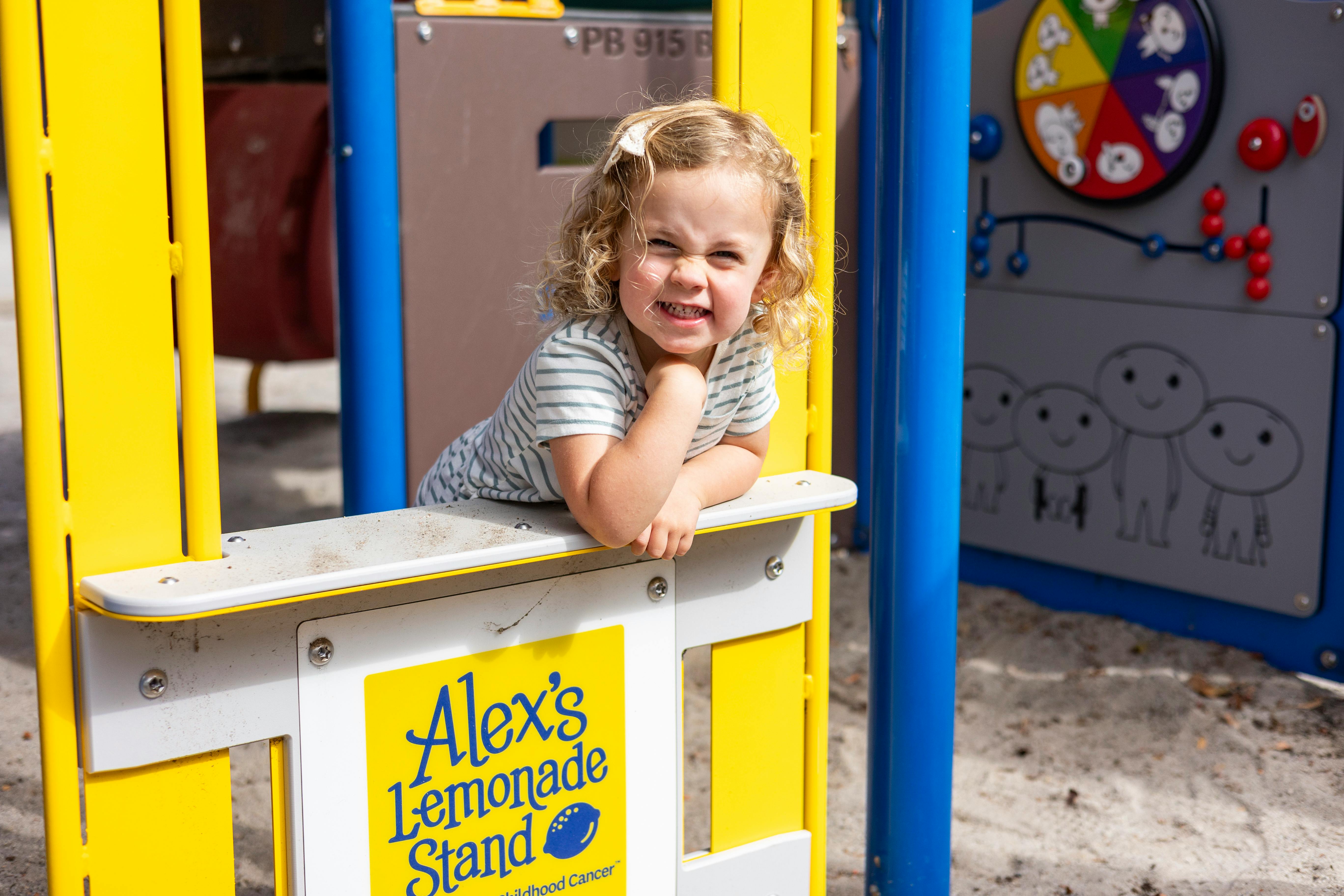 Child leaning on a counter with "Alex's Lemonade Stand" sign