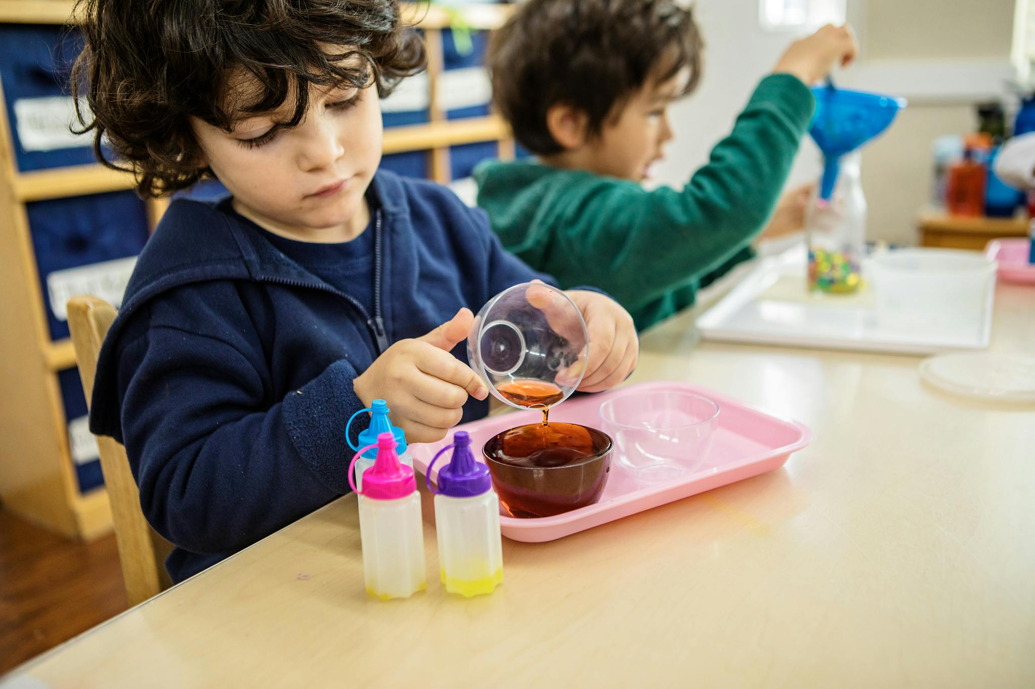 Child pouring liquid from a measuring cup into a bowl on a tray