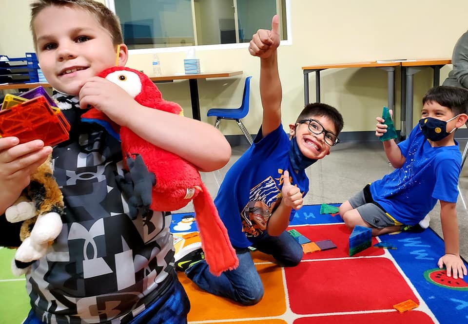 Three children playing on a colorful mat, two giving thumbs up