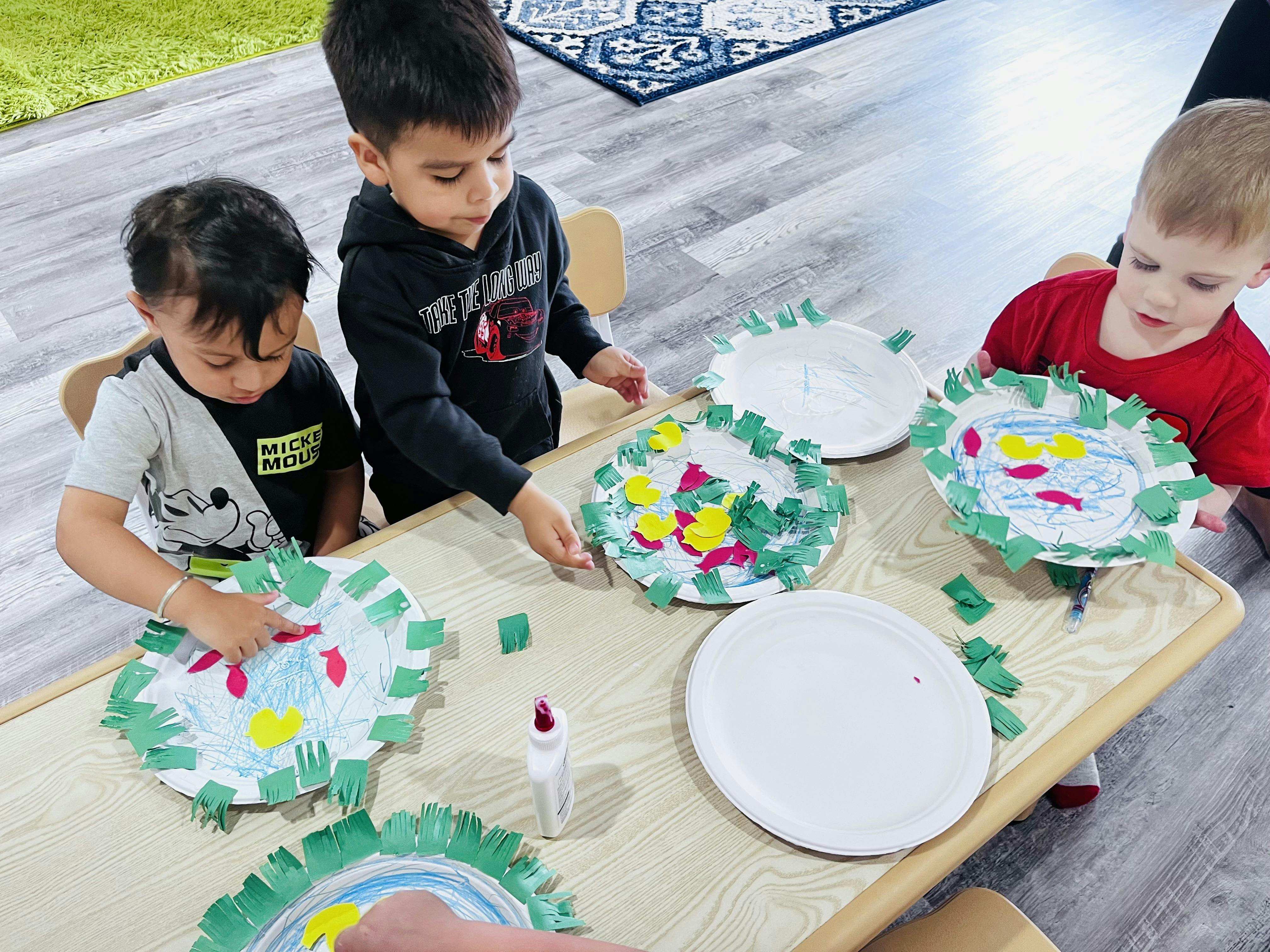 Three children making crafts with paper plates and green paper cutouts
