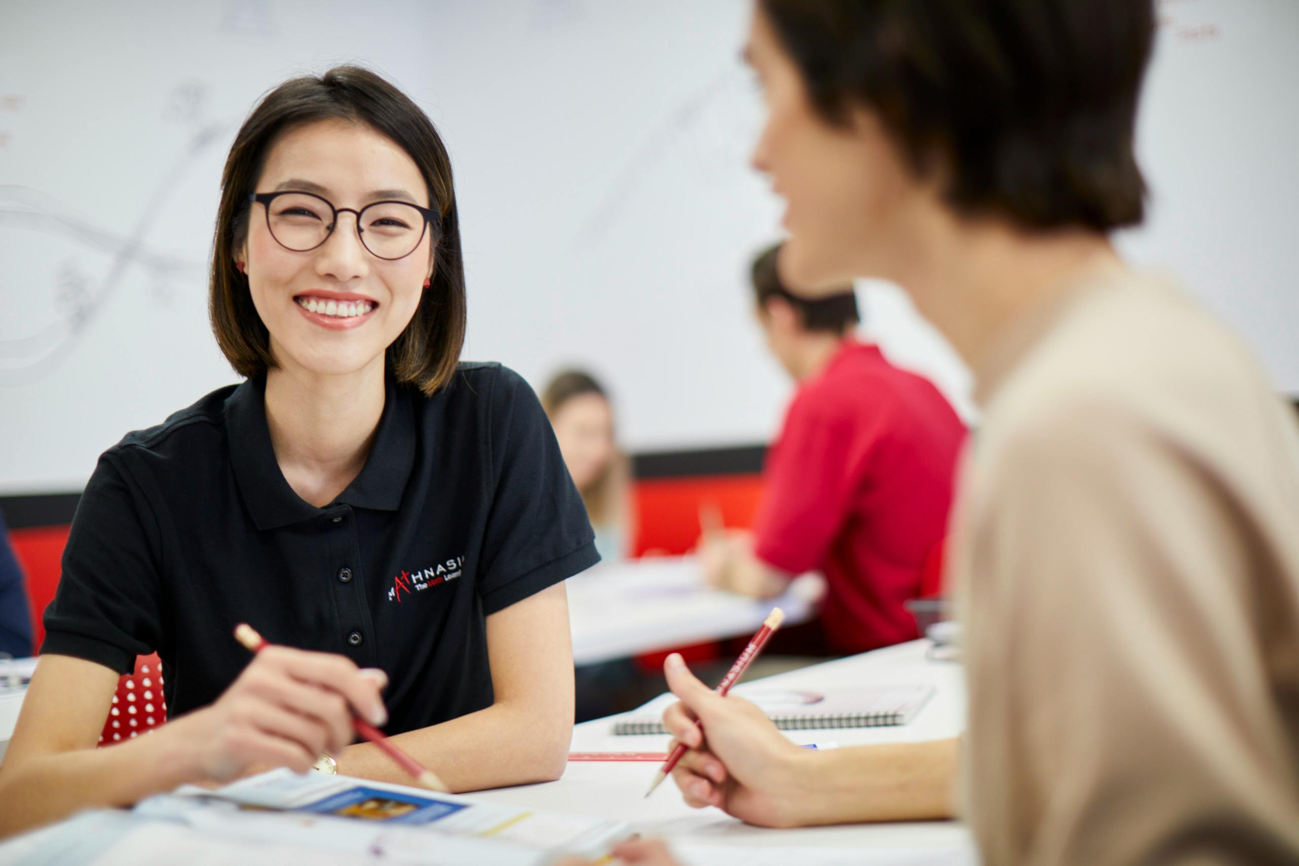 Person with glasses smiling while holding a pencil in a classroom