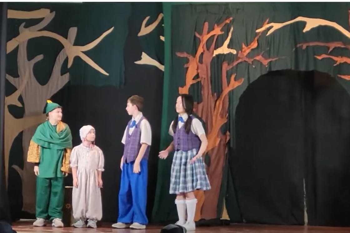 Four children in costumes performing on a stage with a forest backdrop