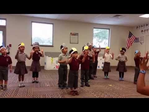 Children in a classroom wearing paper hats and raising their hands