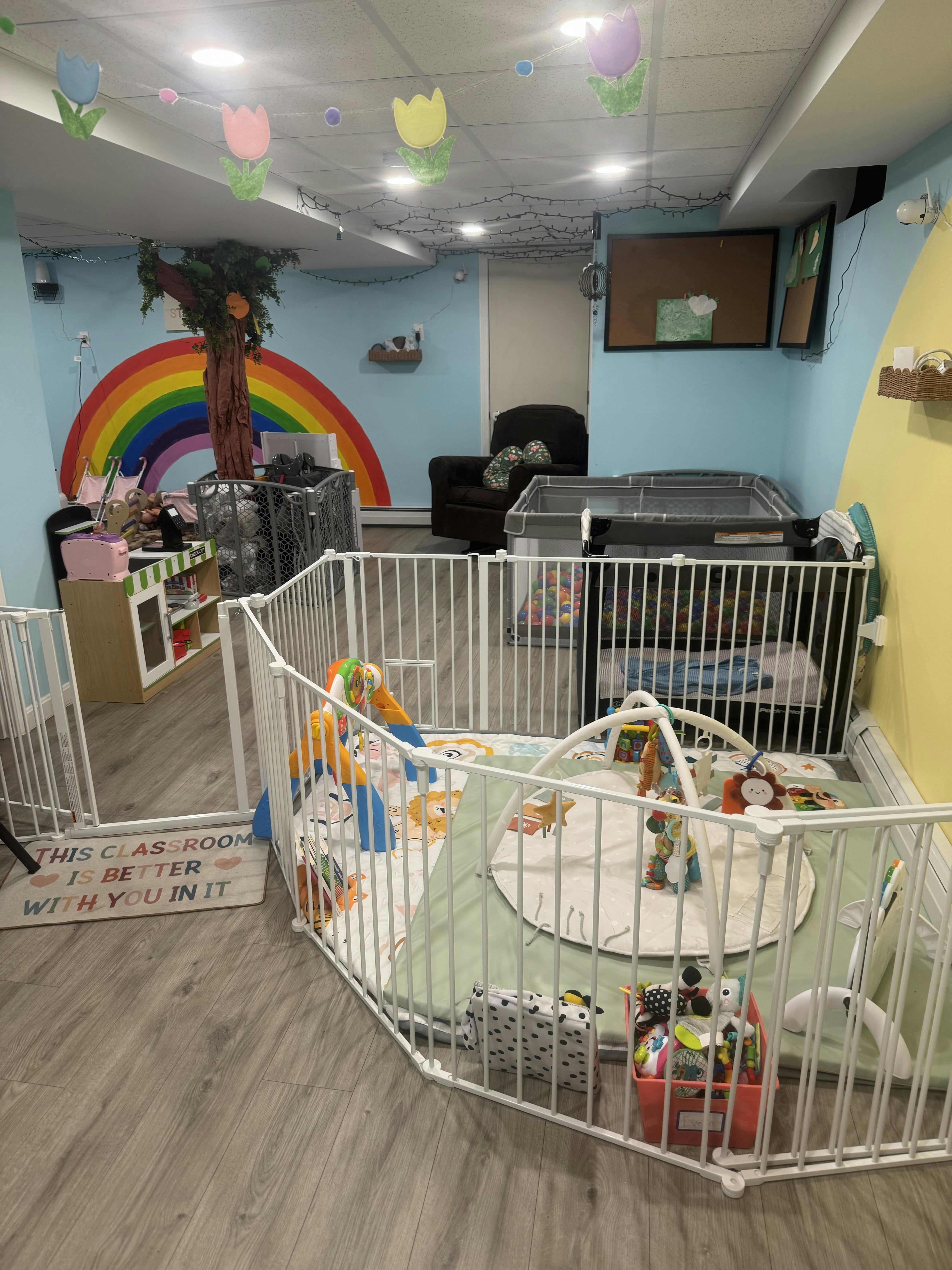 Classroom decorated with rainbow and tree, playpens with toys, and cribs