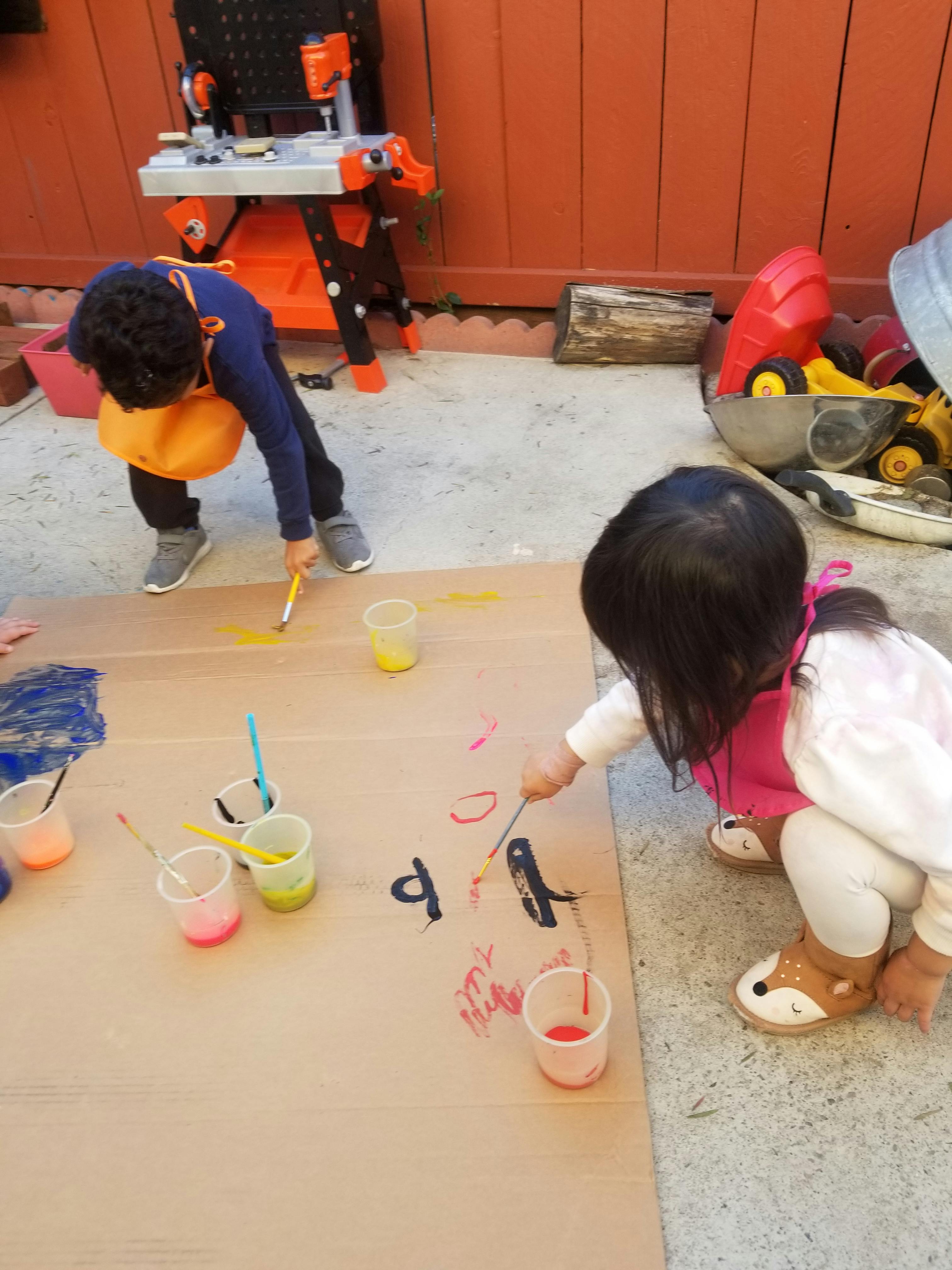 Two children painting on a large piece of cardboard outdoors