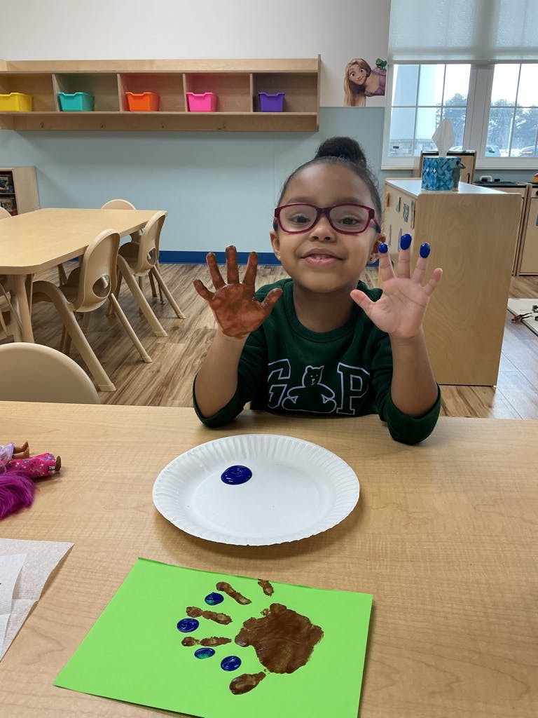 Child in glasses holding up hands covered in brown and blue paint in a classroom