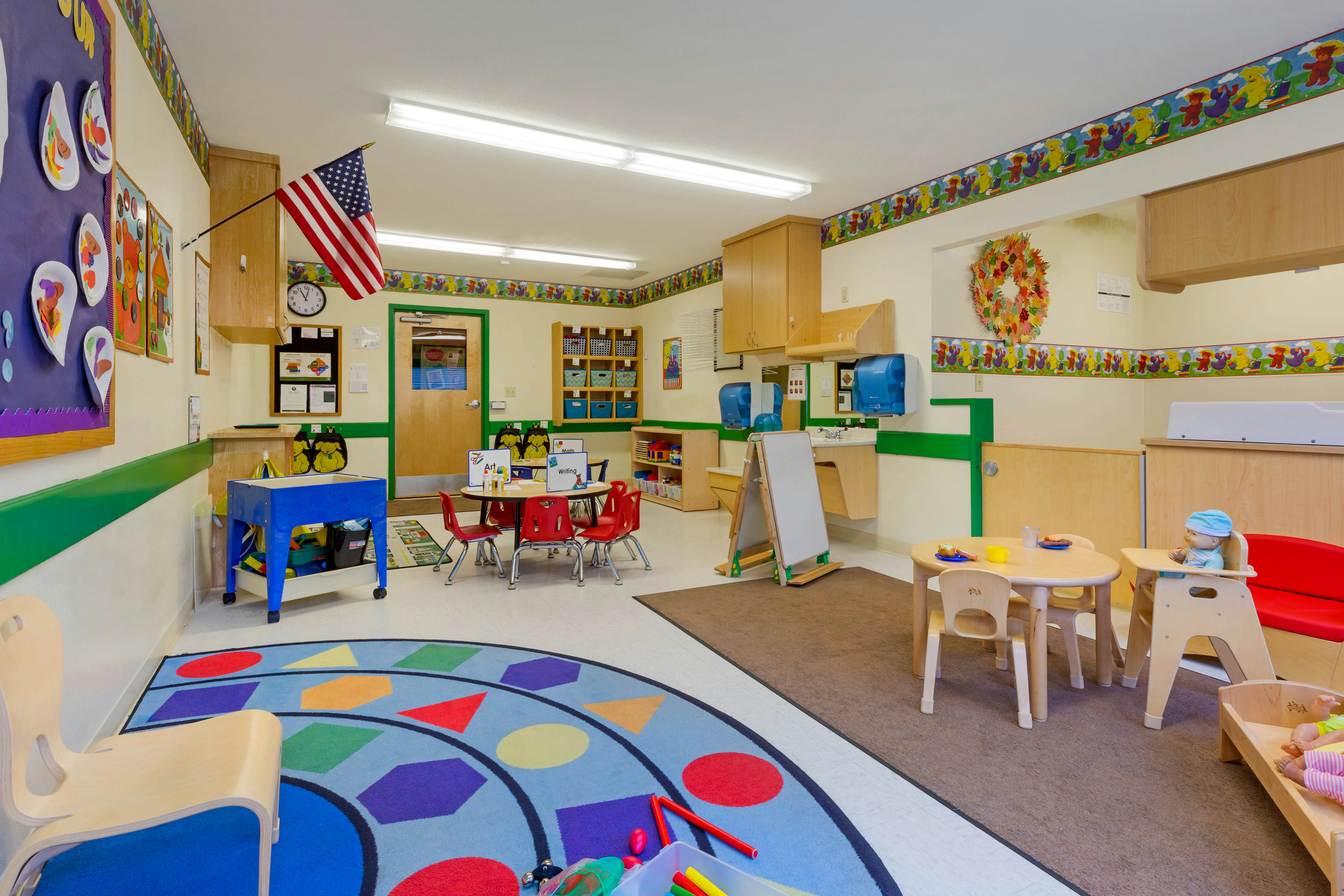 An empty children's classroom with small tables and chairs, and a colorful rug