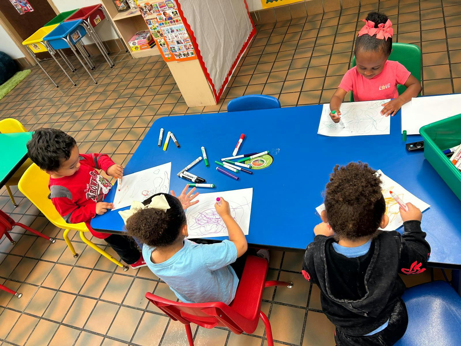 Four children drawing at a blue table in a classroom