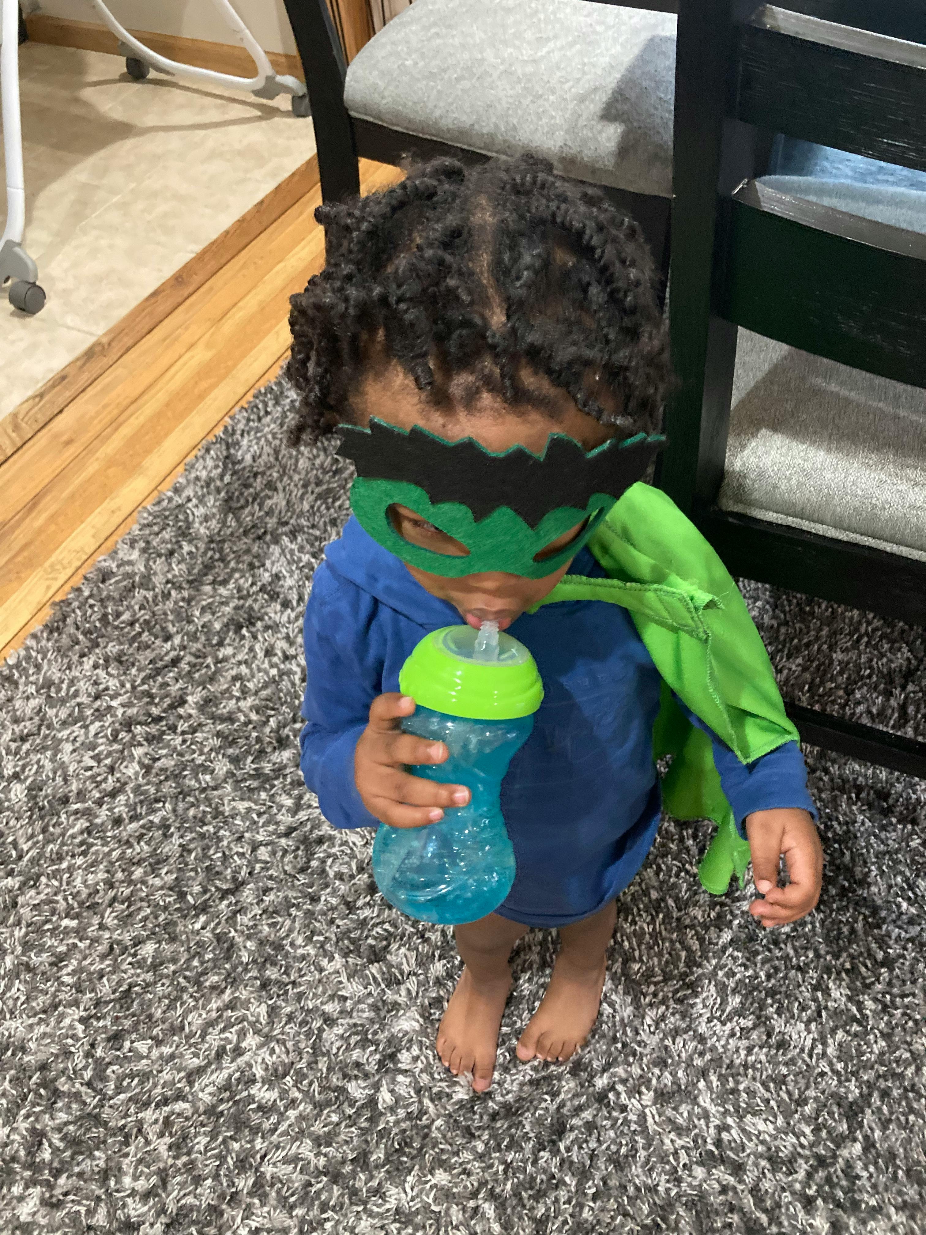 Child wearing a green mask and cape, drinking from a sippy cup