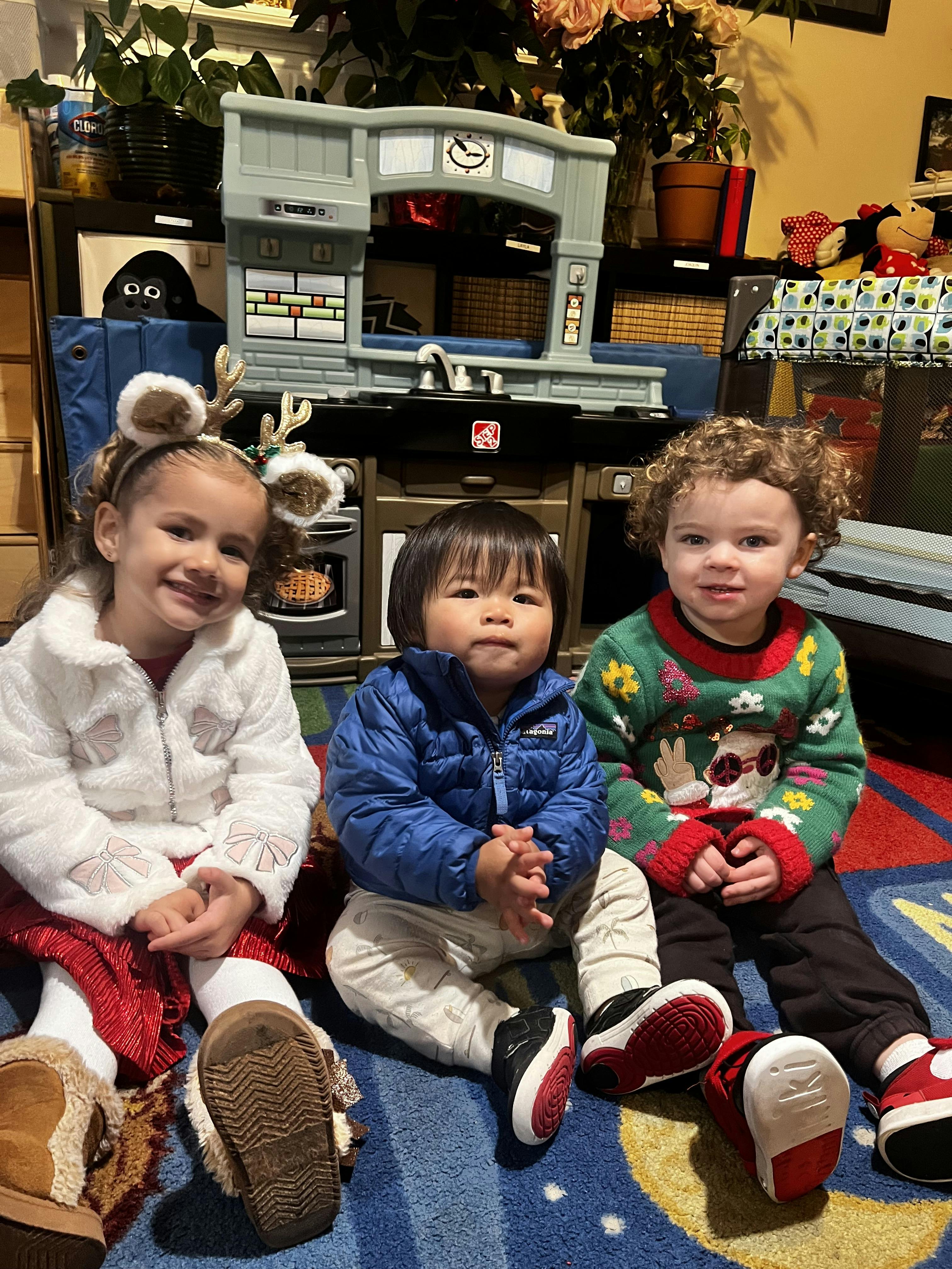 Three children sitting on a rug in front of a play kitchen