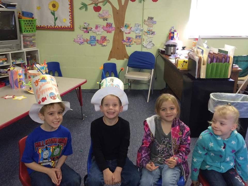 Four children sitting in a classroom, two wearing paper hats