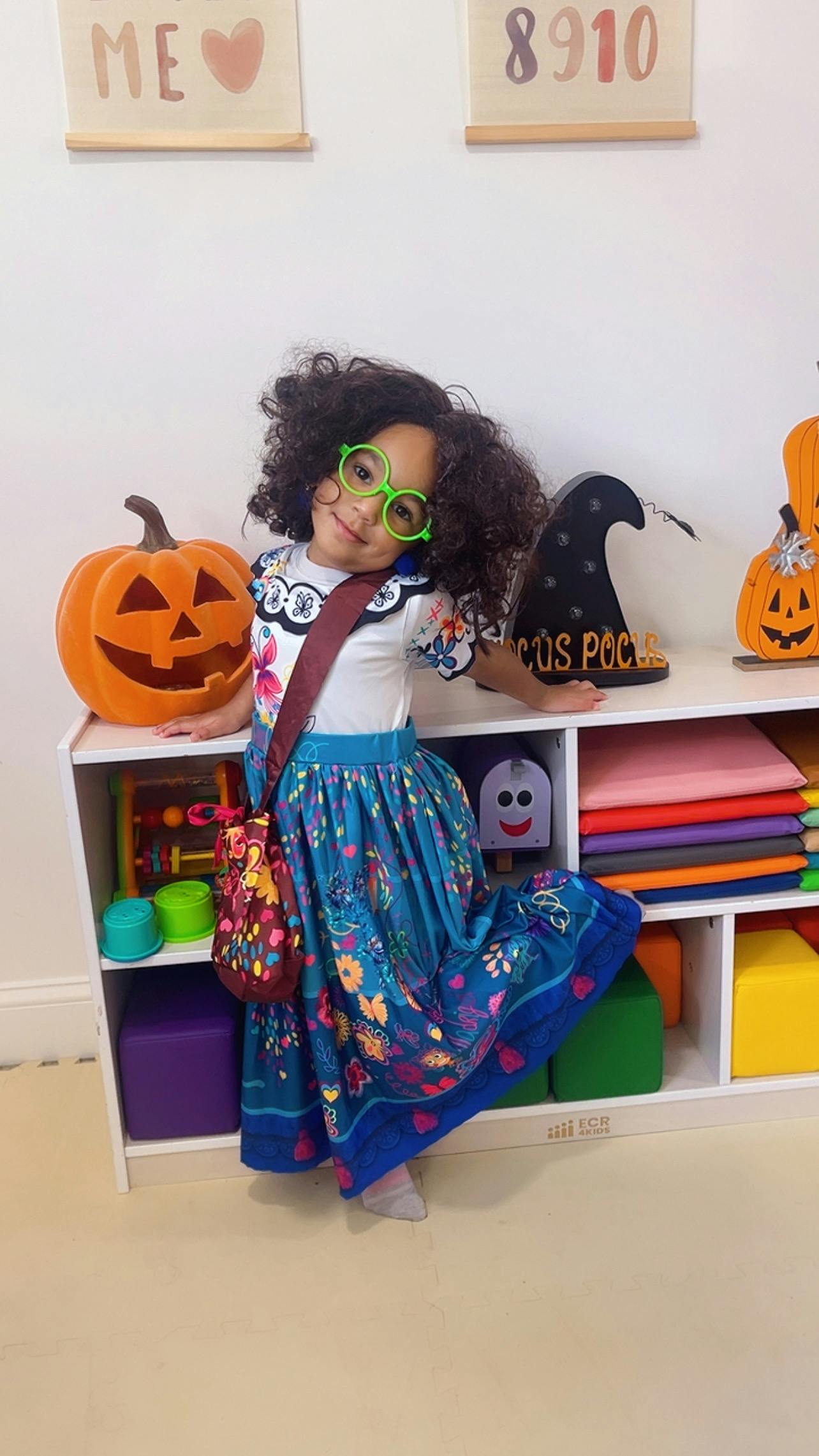 Child in green glasses and a floral skirt standing next to a jack-o'-lantern