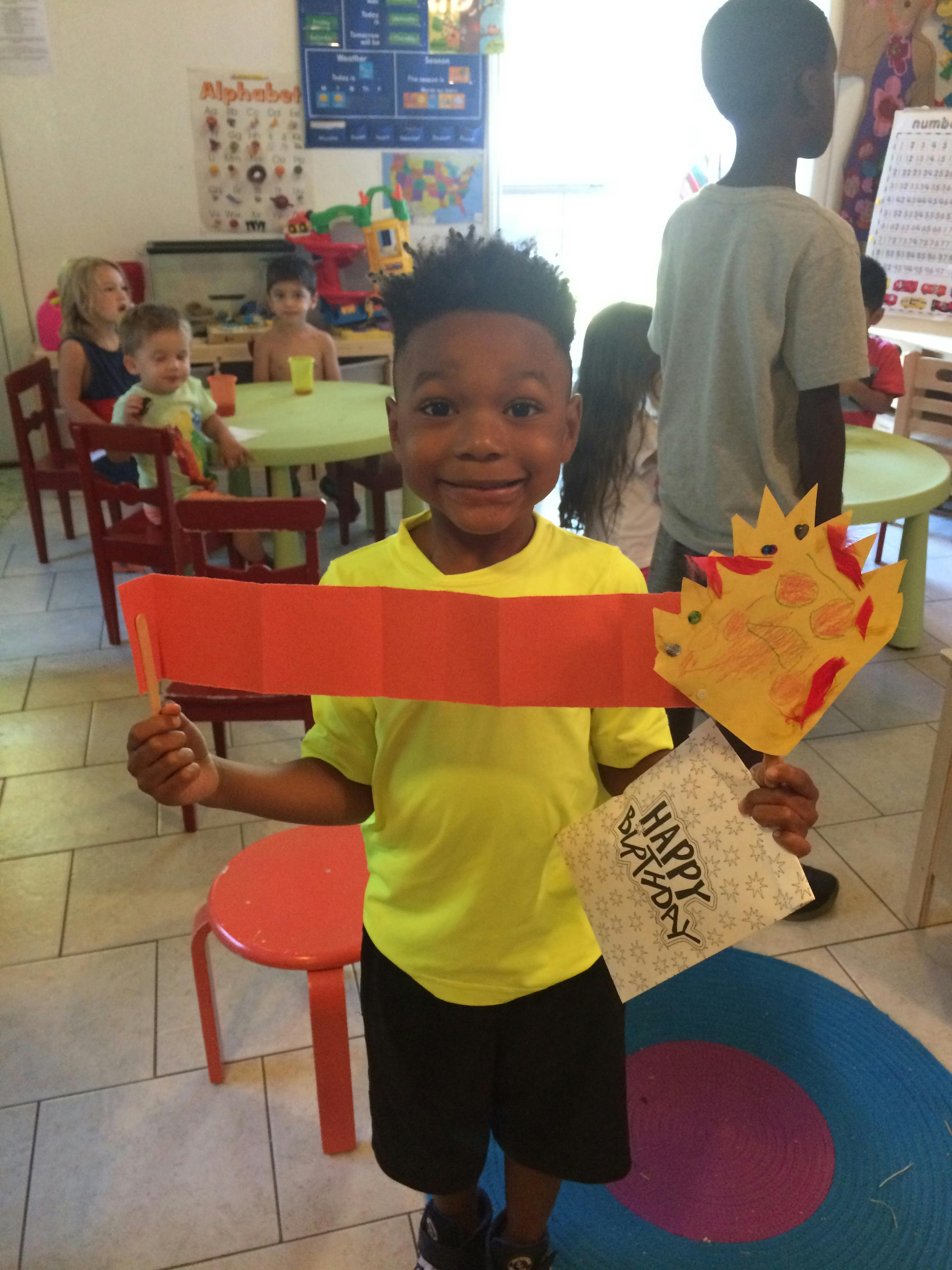 Child holding a paper crown and a red paper strip in a classroom