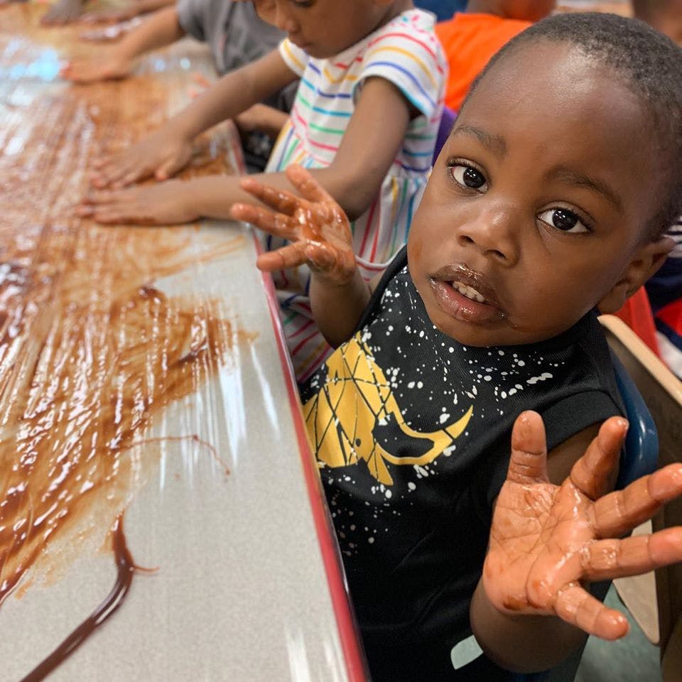 Child with messy hands looking at camera, another child playing with brown substance