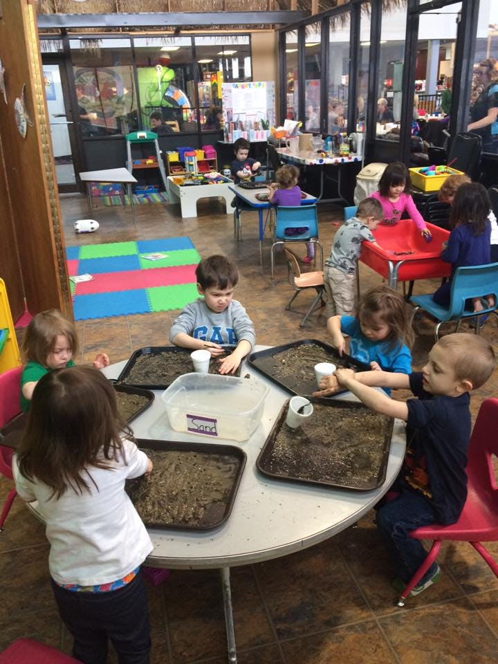 Children playing with trays of dirt and cups at a table in a classroom