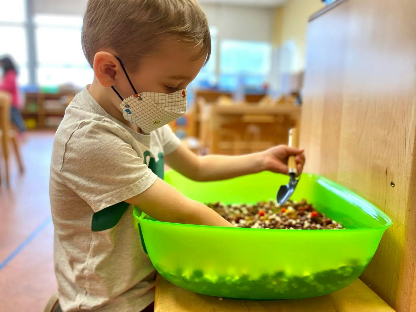 Child in a mask playing with a shovel in a green bin