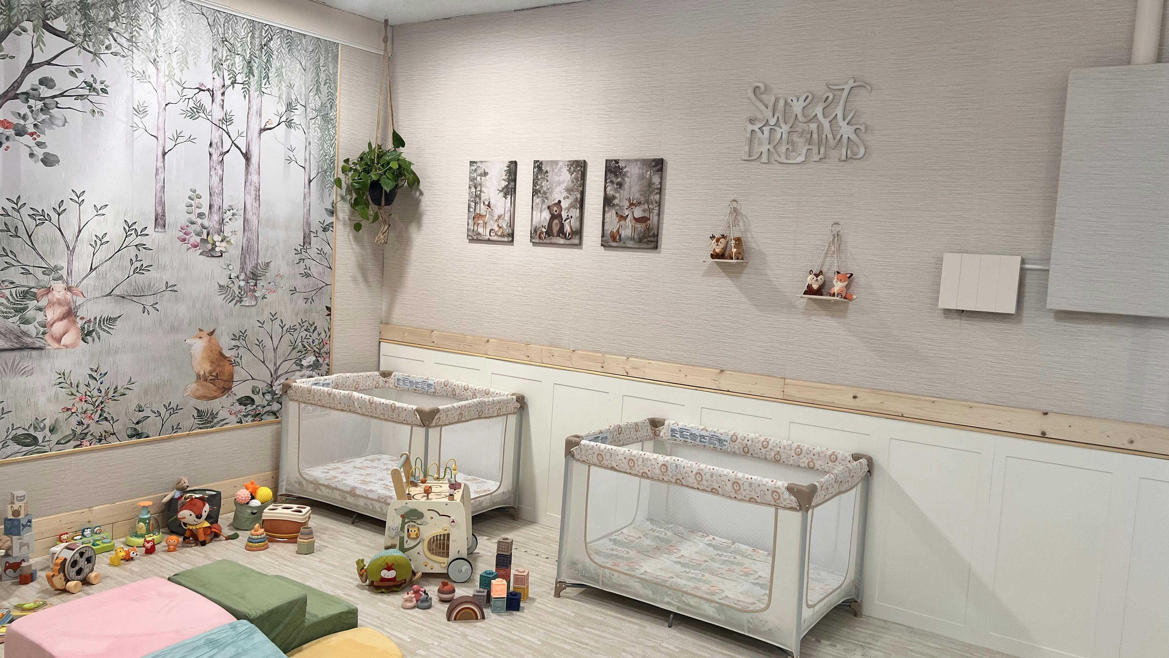 Two cribs in a room with toys on the floor and a forest mural on the wall