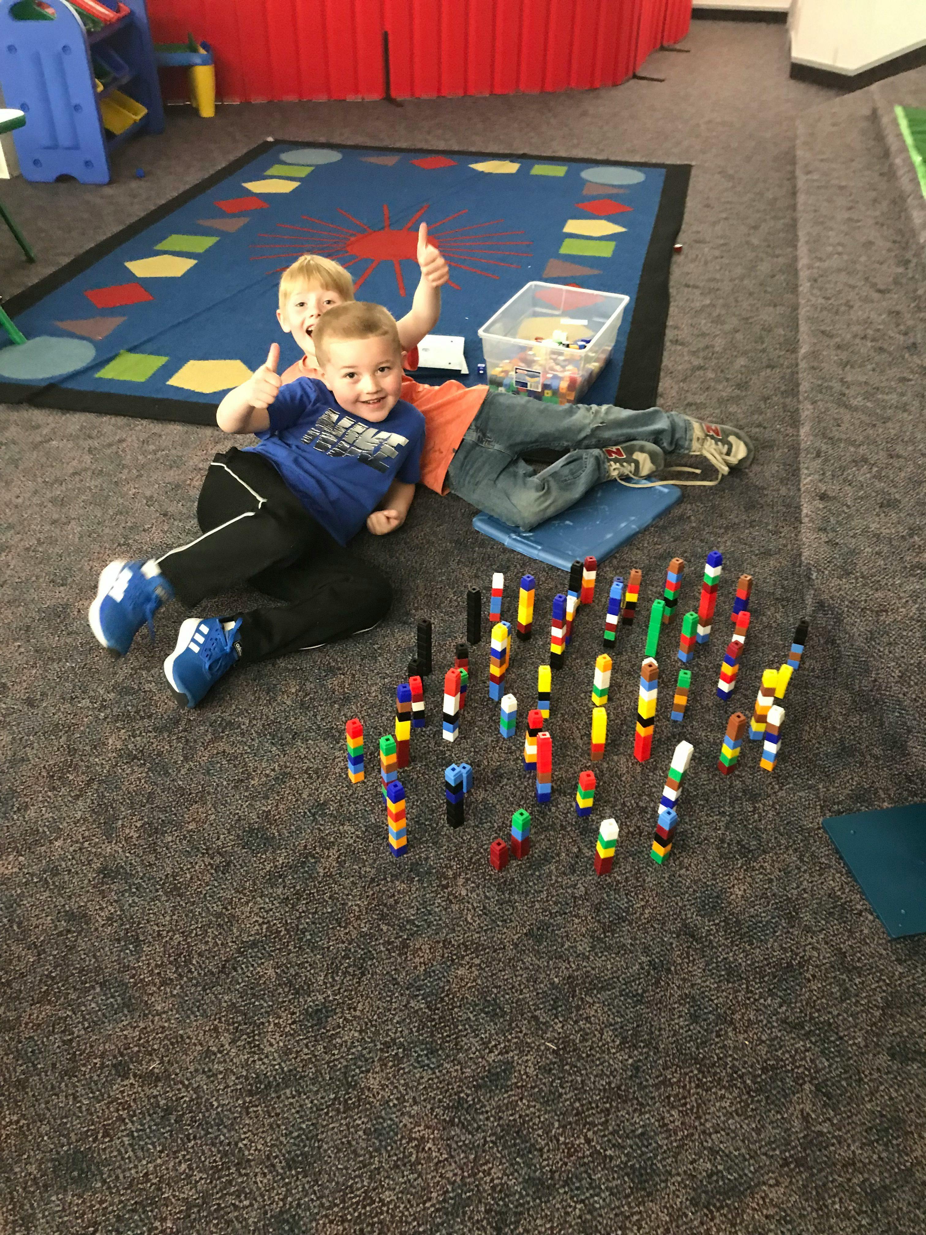 Two children giving thumbs up while lying on the floor with colorful blocks