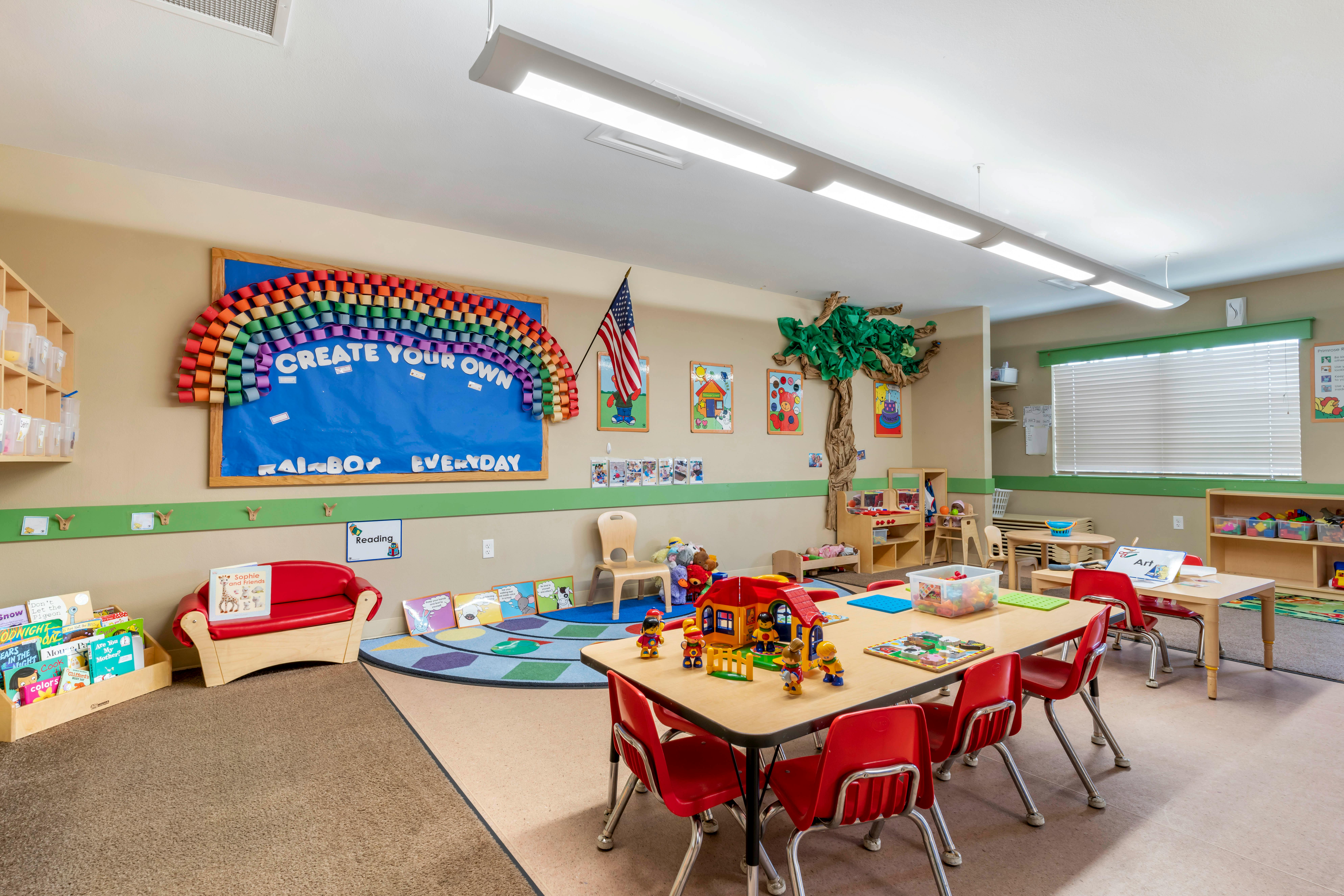 A colorful preschool classroom with tables, chairs, toys, and a large rainbow bulletin board