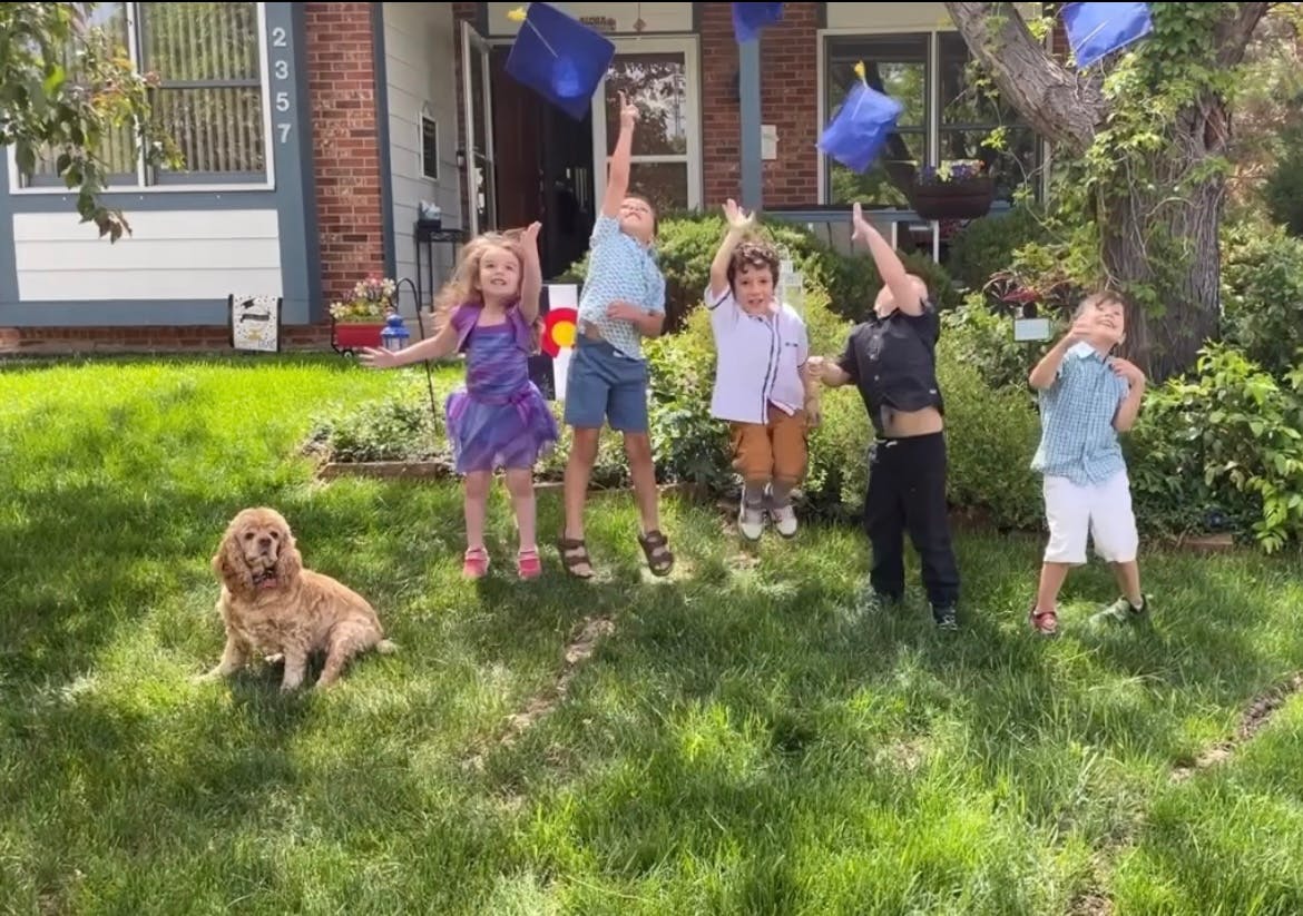 Five children jumping in a yard, throwing graduation caps into the air, with a dog sitting nearby