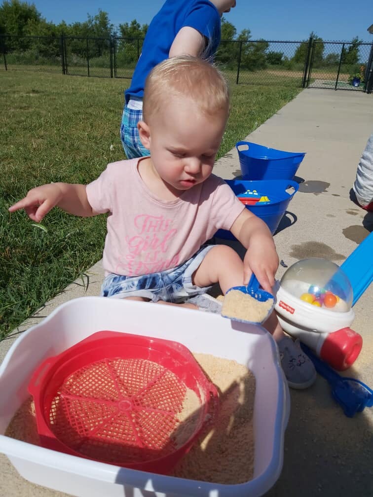 Child playing with sand and a red sieve in a white bin